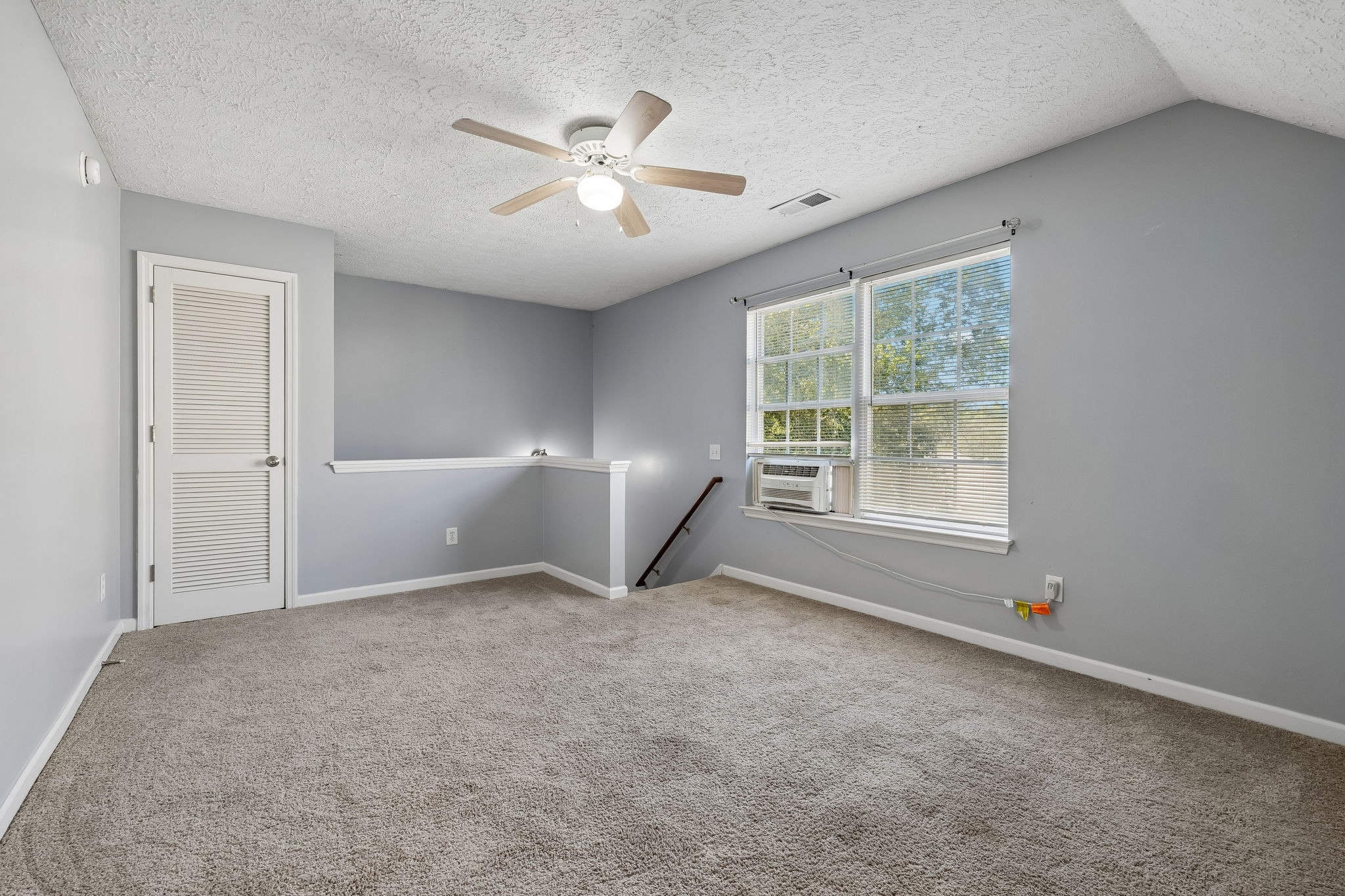 Empty carpeted room with a ceiling fan, louvered door, and windows with blinds.