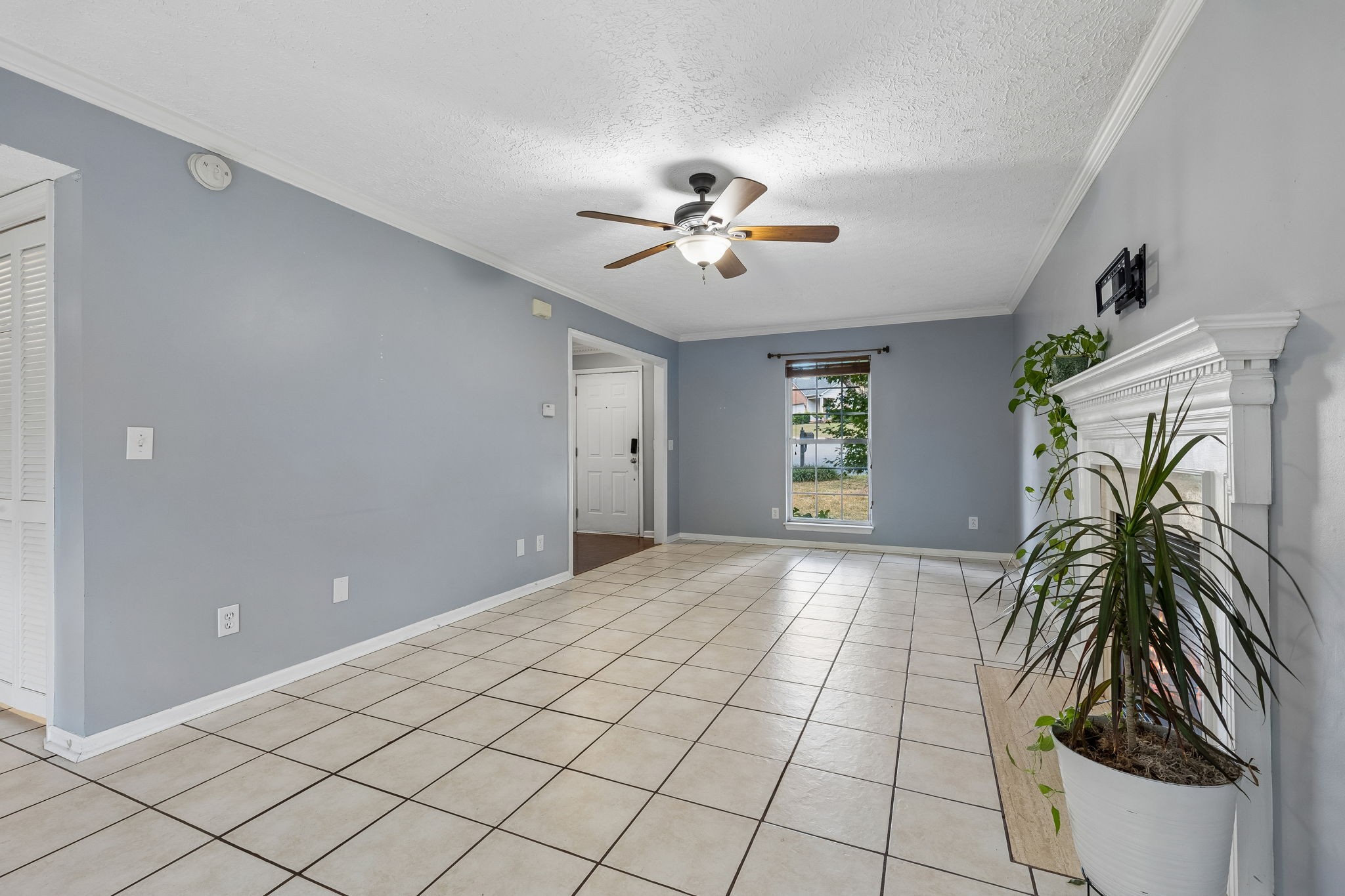 Empty living room with gray walls, large tiled floor, white fireplace, ceiling fan, and potted plant.