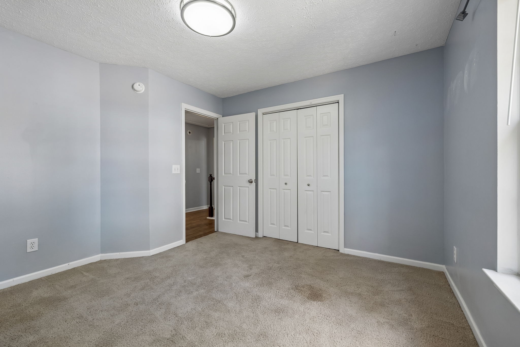 Empty bedroom with beige carpet, pale blue-gray walls, and white double closet doors.