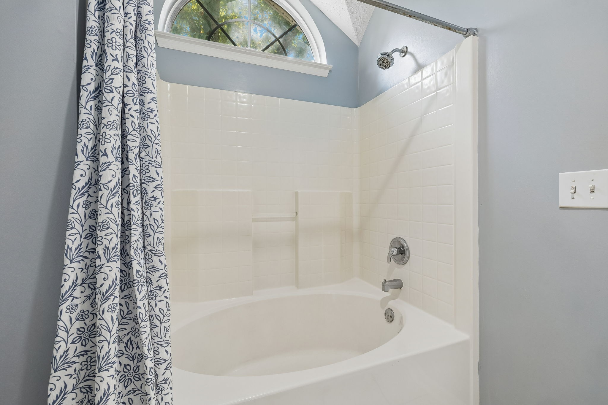 Bathroom tub and tiled shower beneath an arched window, with a blue-and-white floral curtain.