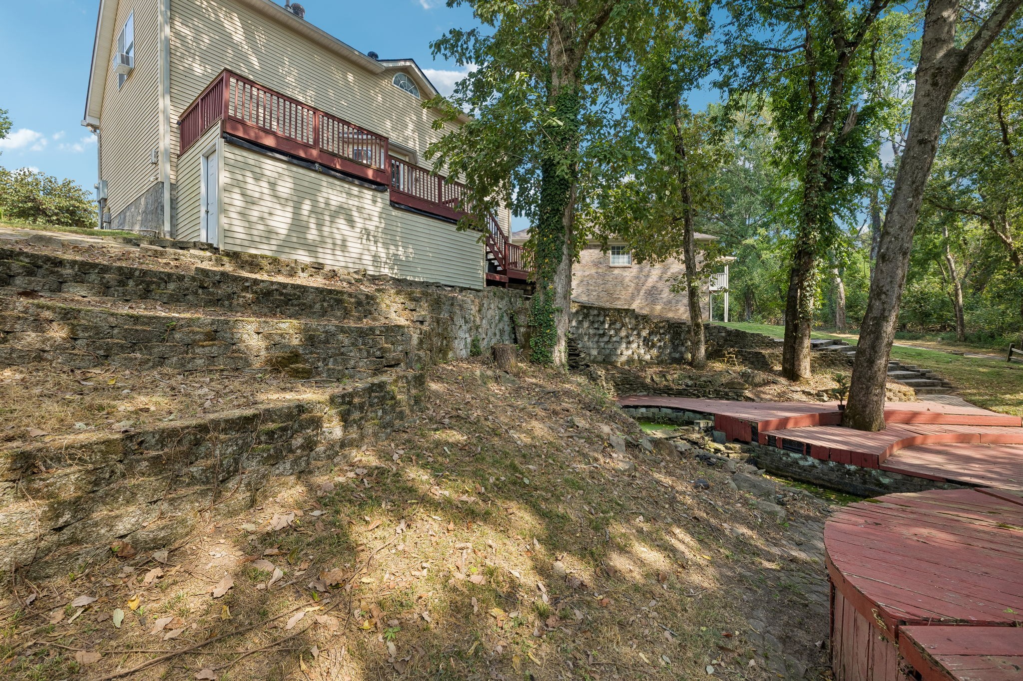 Home’s rear deck overlooks terraced stone retaining walls and wooden walkways around mature trees.