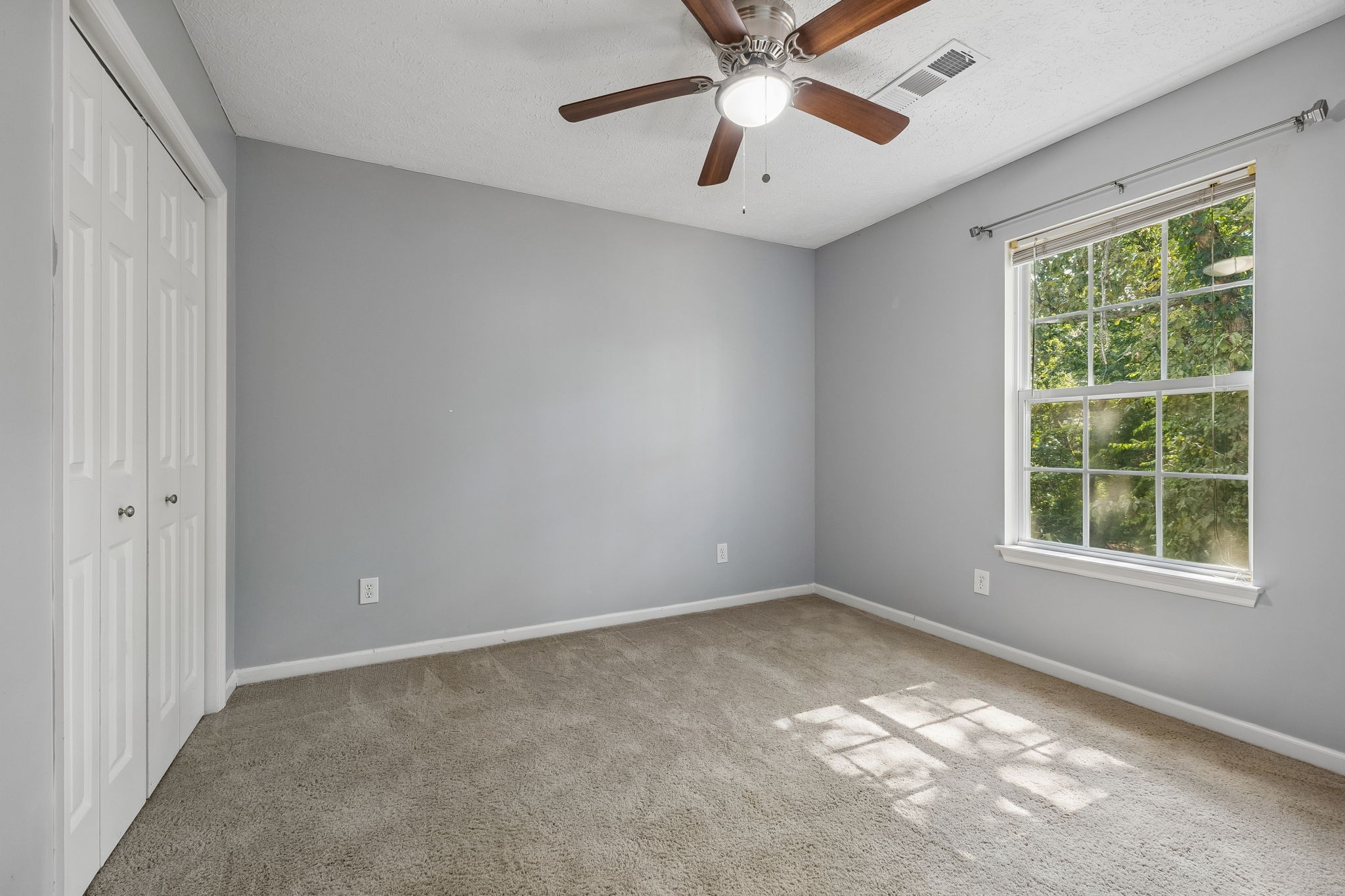 Empty bedroom with gray walls, beige carpet, a double-door closet, and a sunny window overlooking trees.