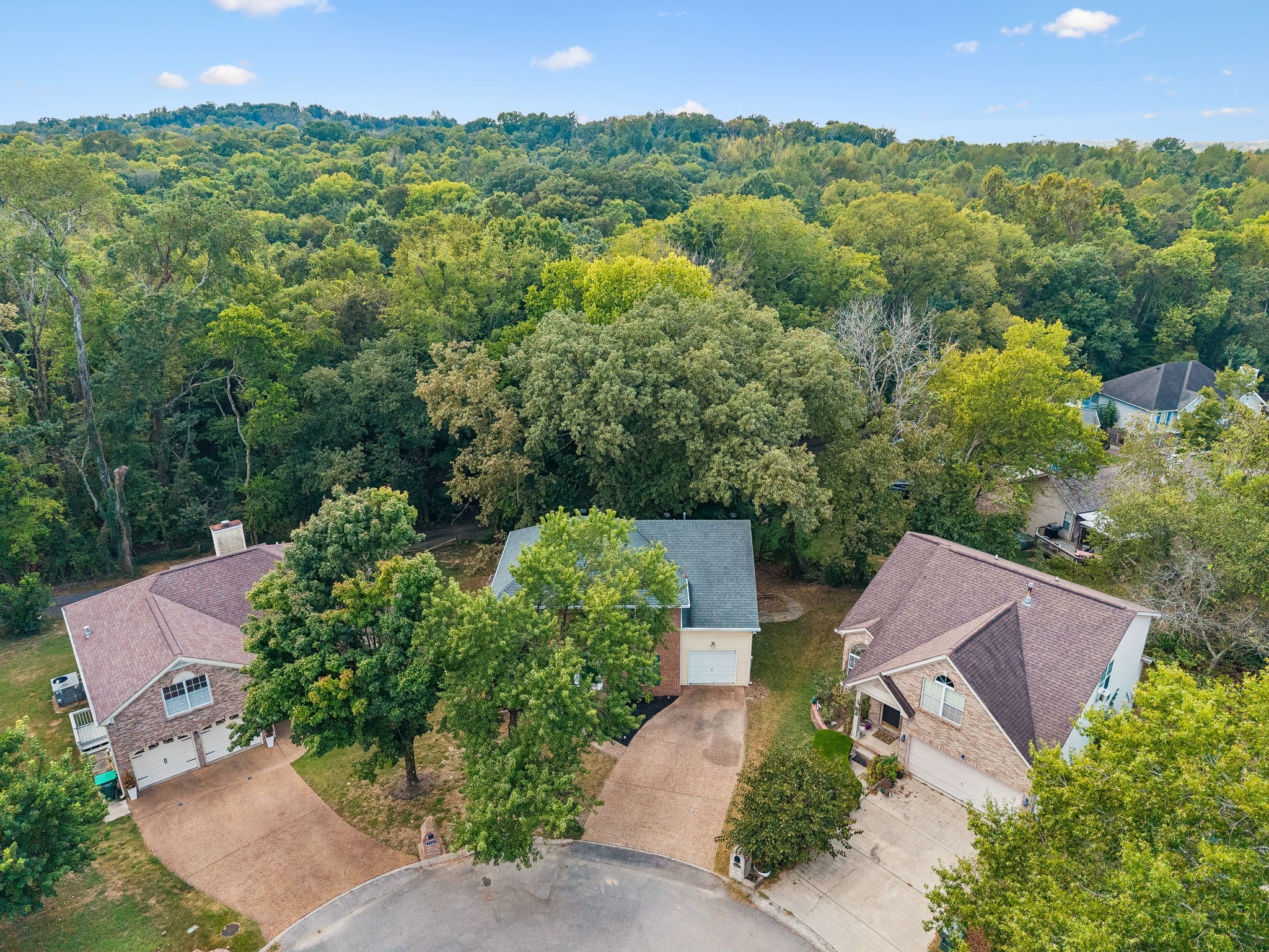 Aerial view of a brick home with a gray roof, driveway, and mature trees behind it.