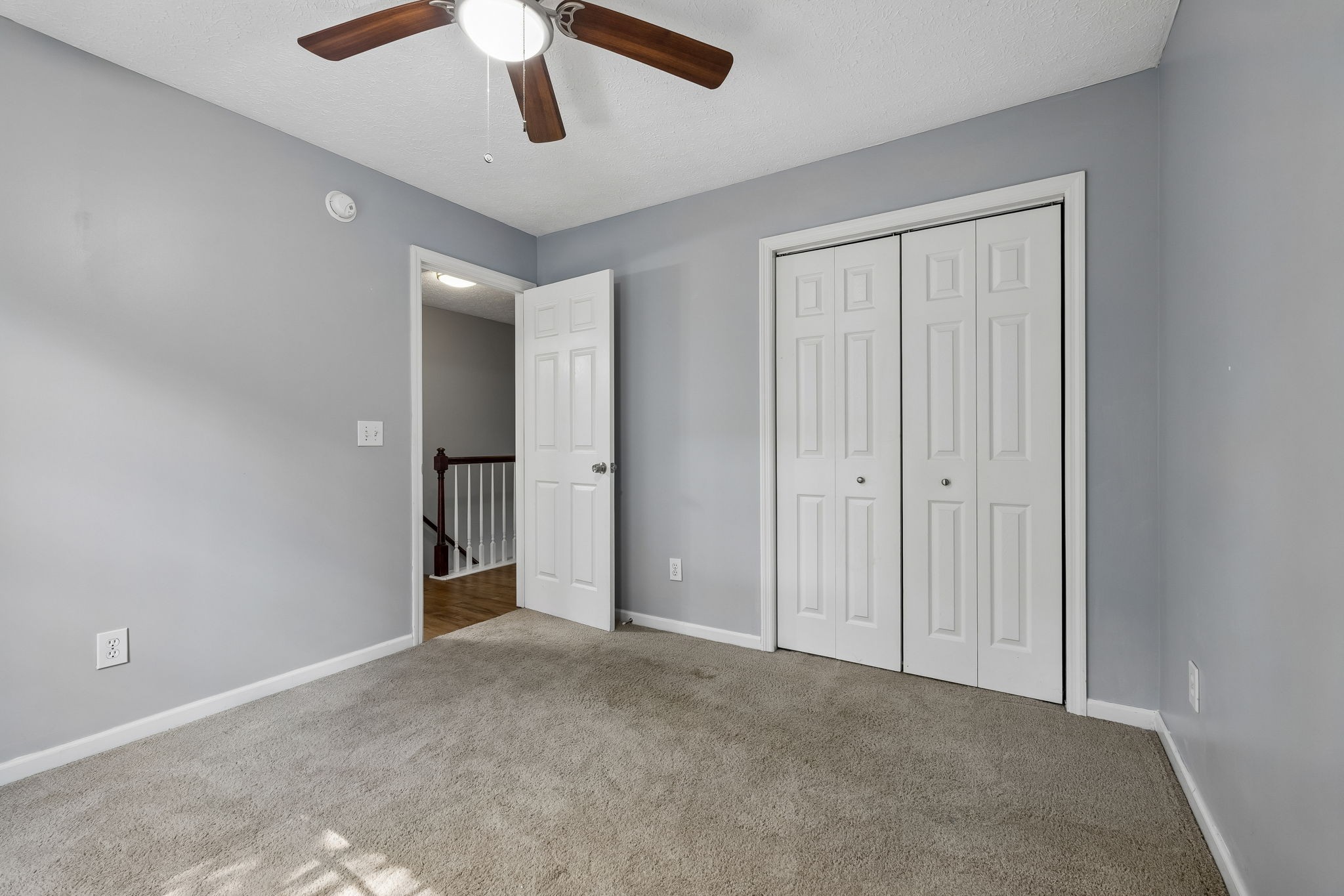 Empty gray bedroom with beige carpet, ceiling fan, white closet doors, and an open doorway to the landing.
