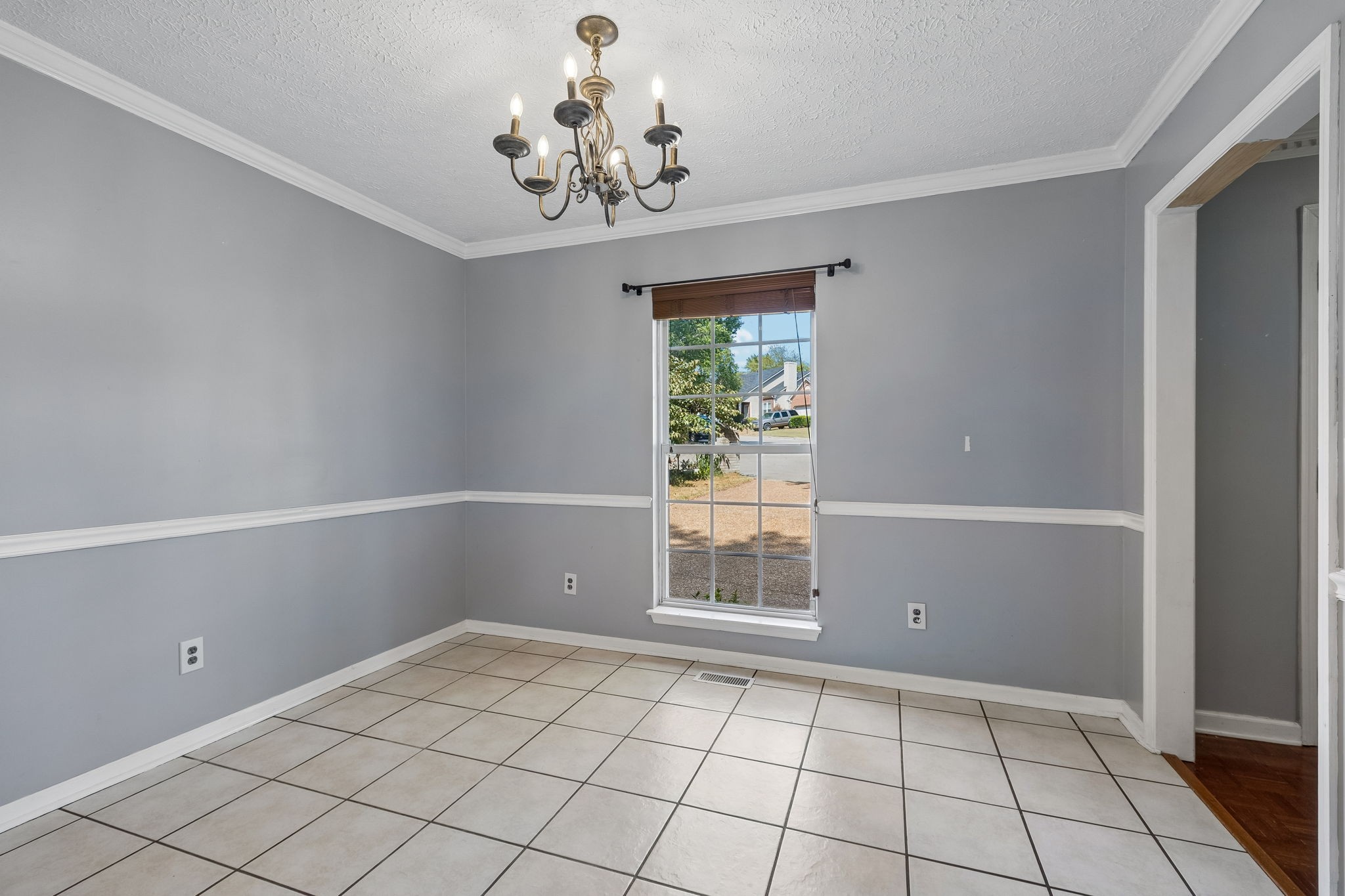 Empty dining room with gray walls, tiled floor, brass chandelier, and a street-facing window.