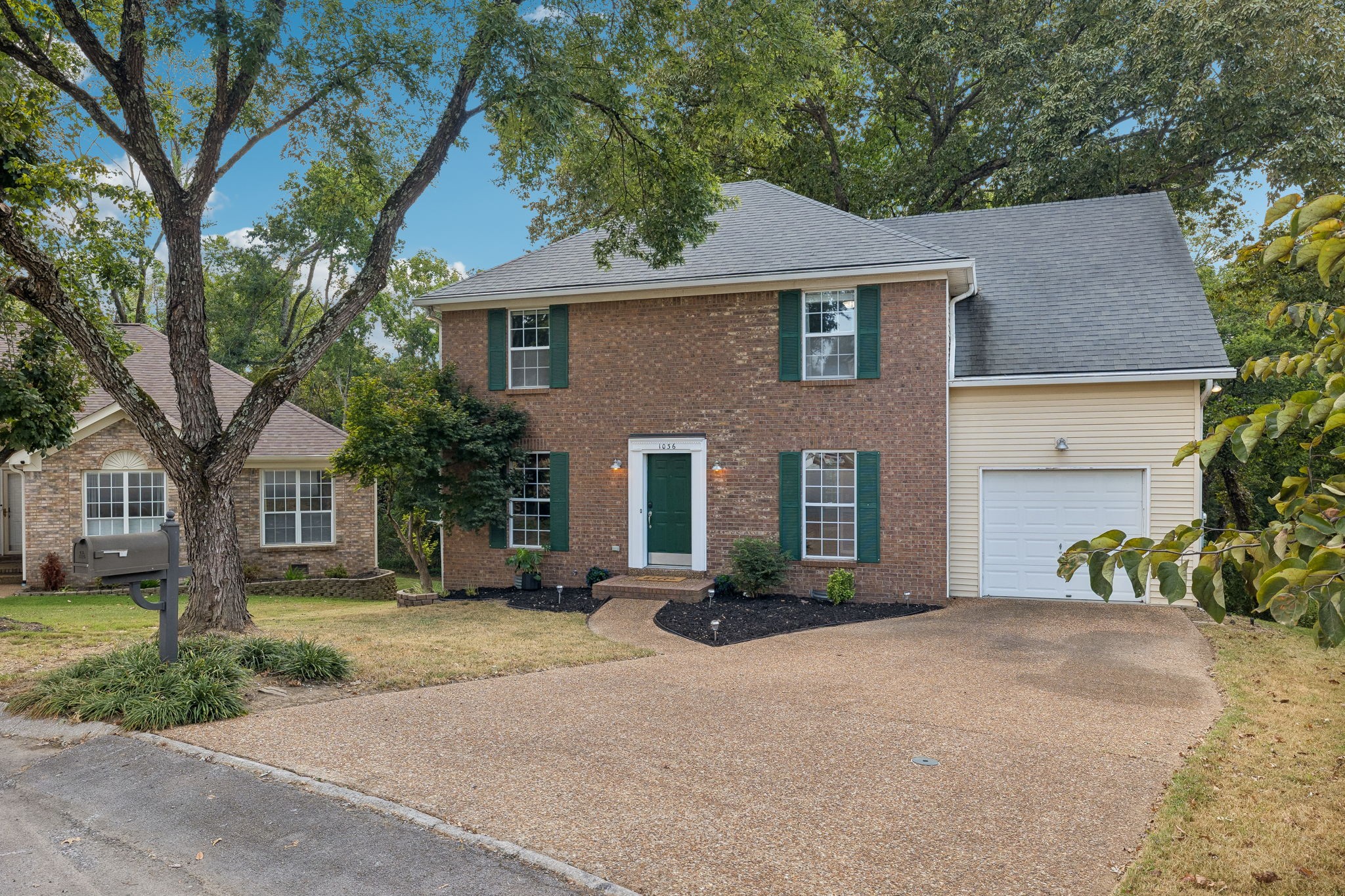 Two-story brick house with green shutters, an attached garage, and a wide driveway beneath mature trees.