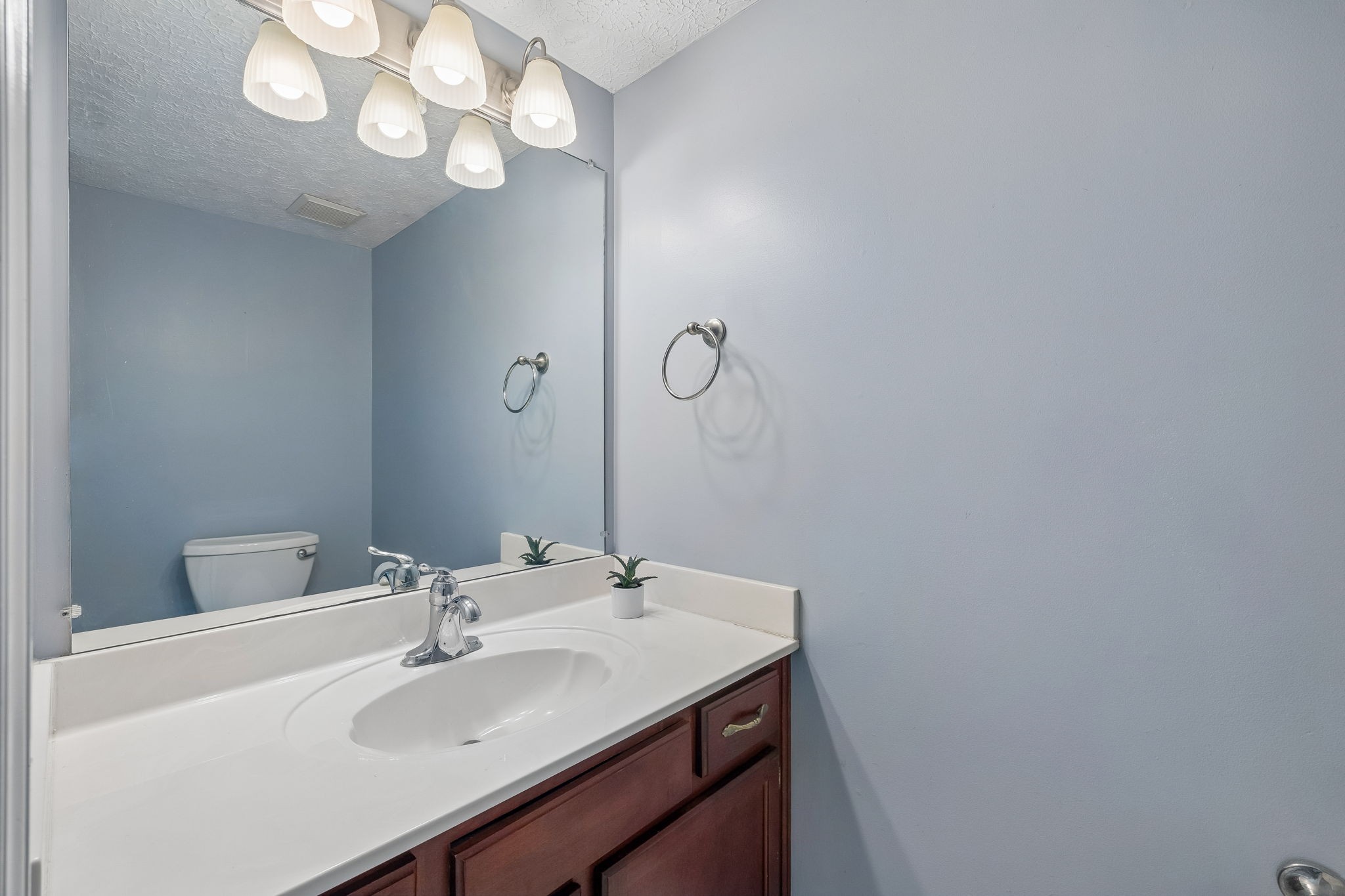Bathroom vanity with white countertop, oval sink, dark wood cabinetry, and a large mirror reflecting the toilet.