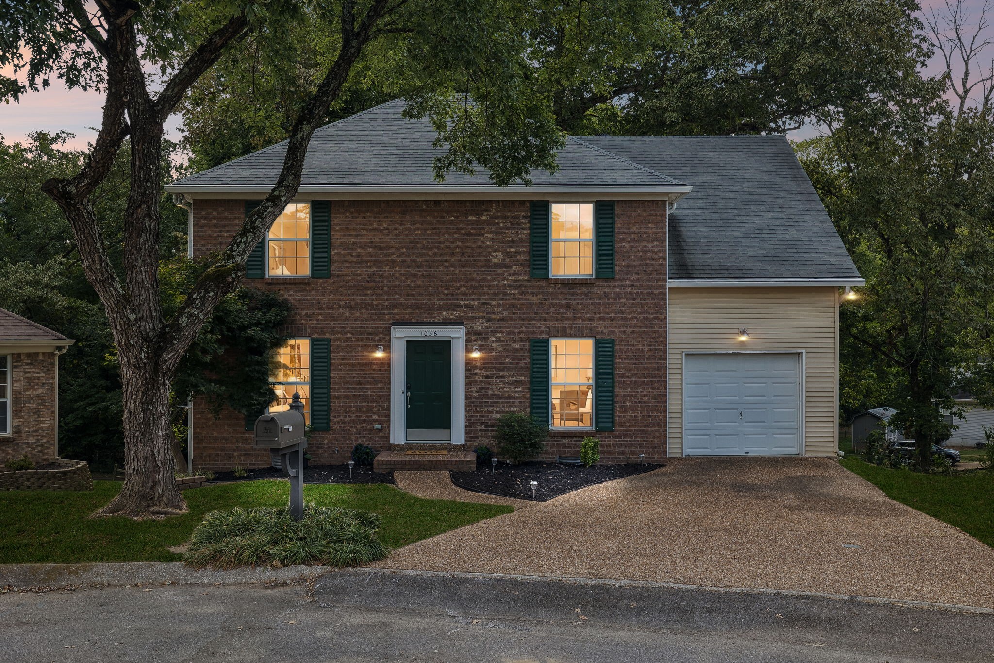Two-story brick house at dusk with a one-car garage, warmly lit windows, and a large front-yard tree.