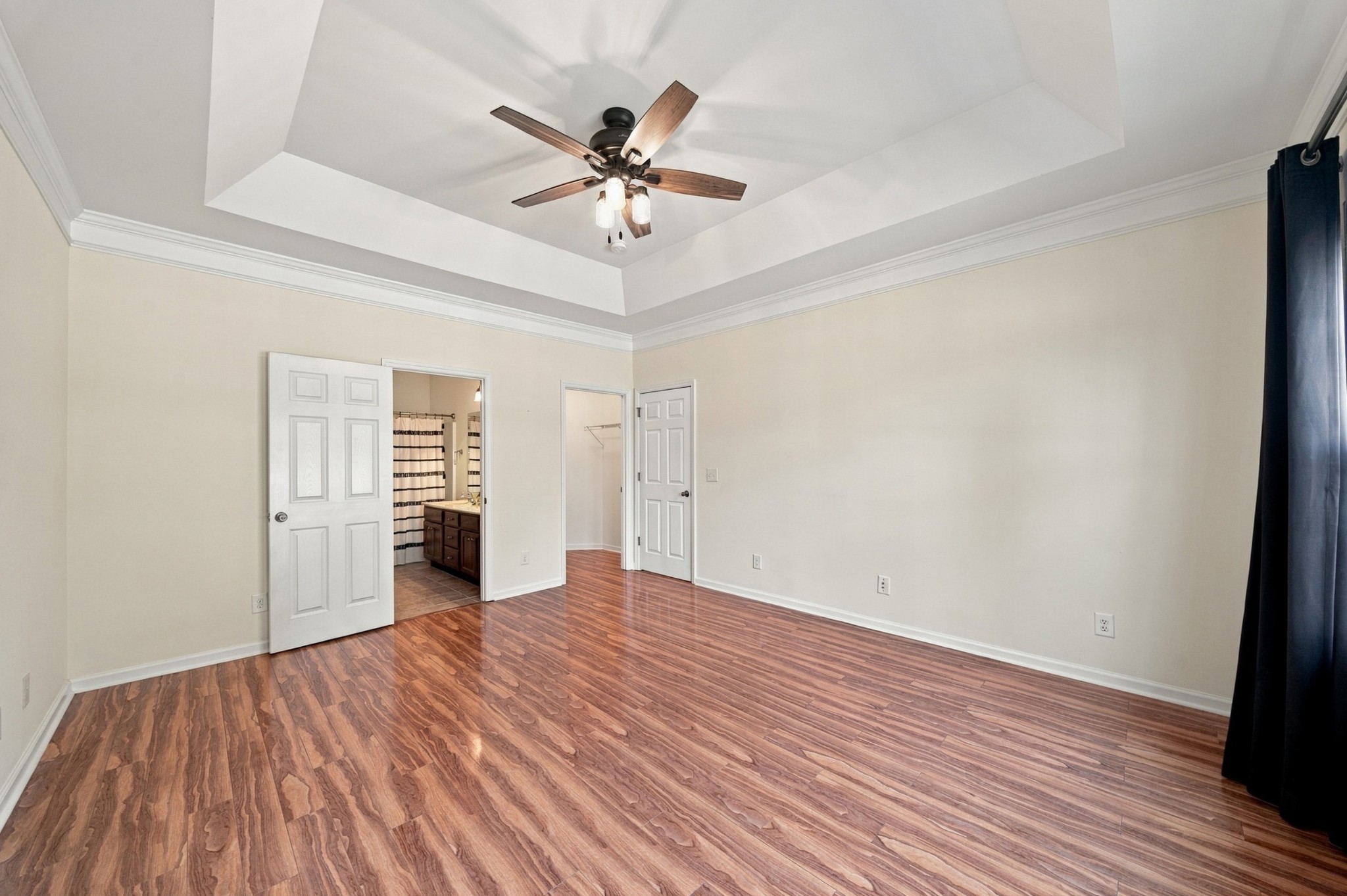 Empty bedroom with wood-look floors, a tray ceiling, and adjoining bathroom and closet doors.