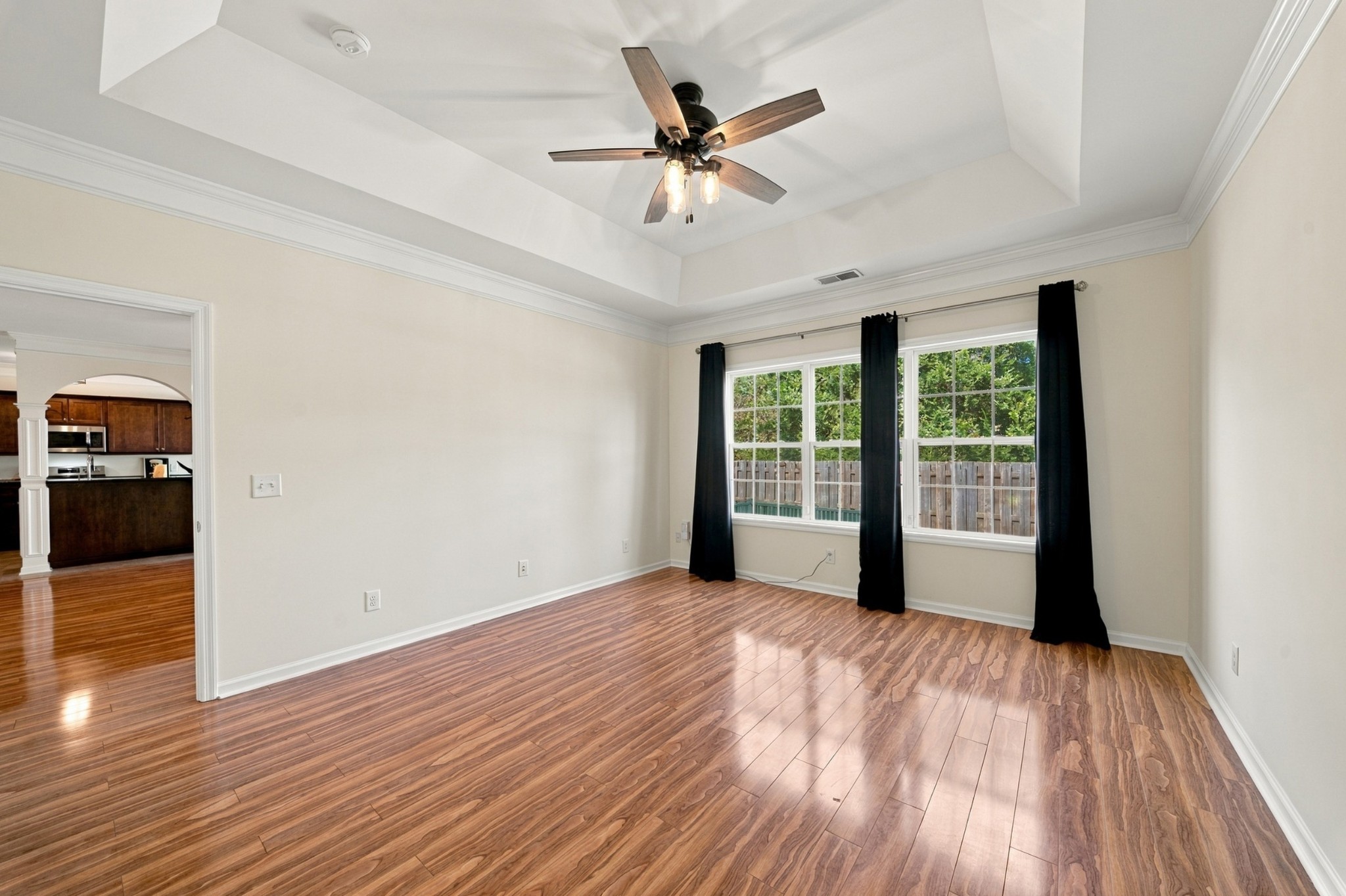 Vacant living room with wood-look floors, a tray ceiling, and curtained windows overlooking a fenced yard.