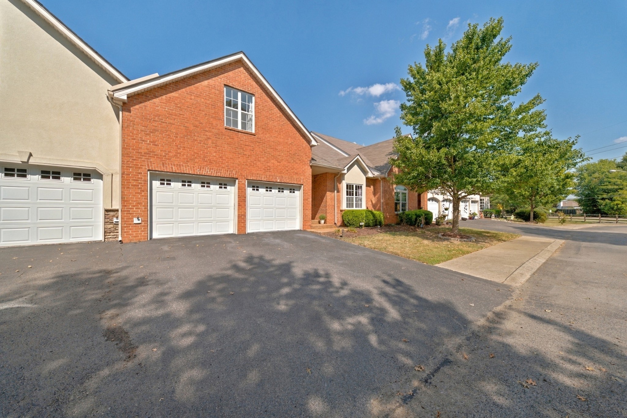 Brick townhouse exterior with white garage doors, a paved driveway, and a large leafy tree.