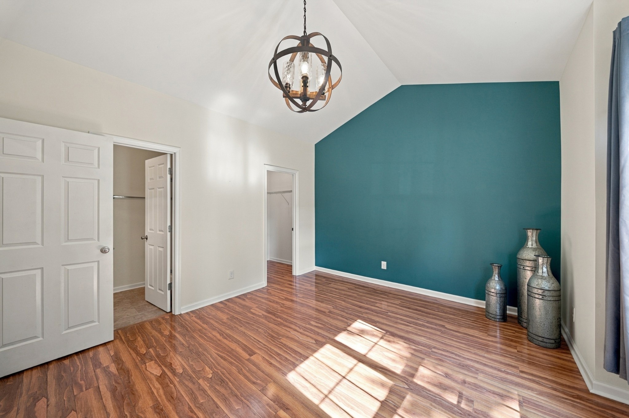 Empty bedroom with a teal accent wall, wood-look floors, and sunlight streaming across the room.