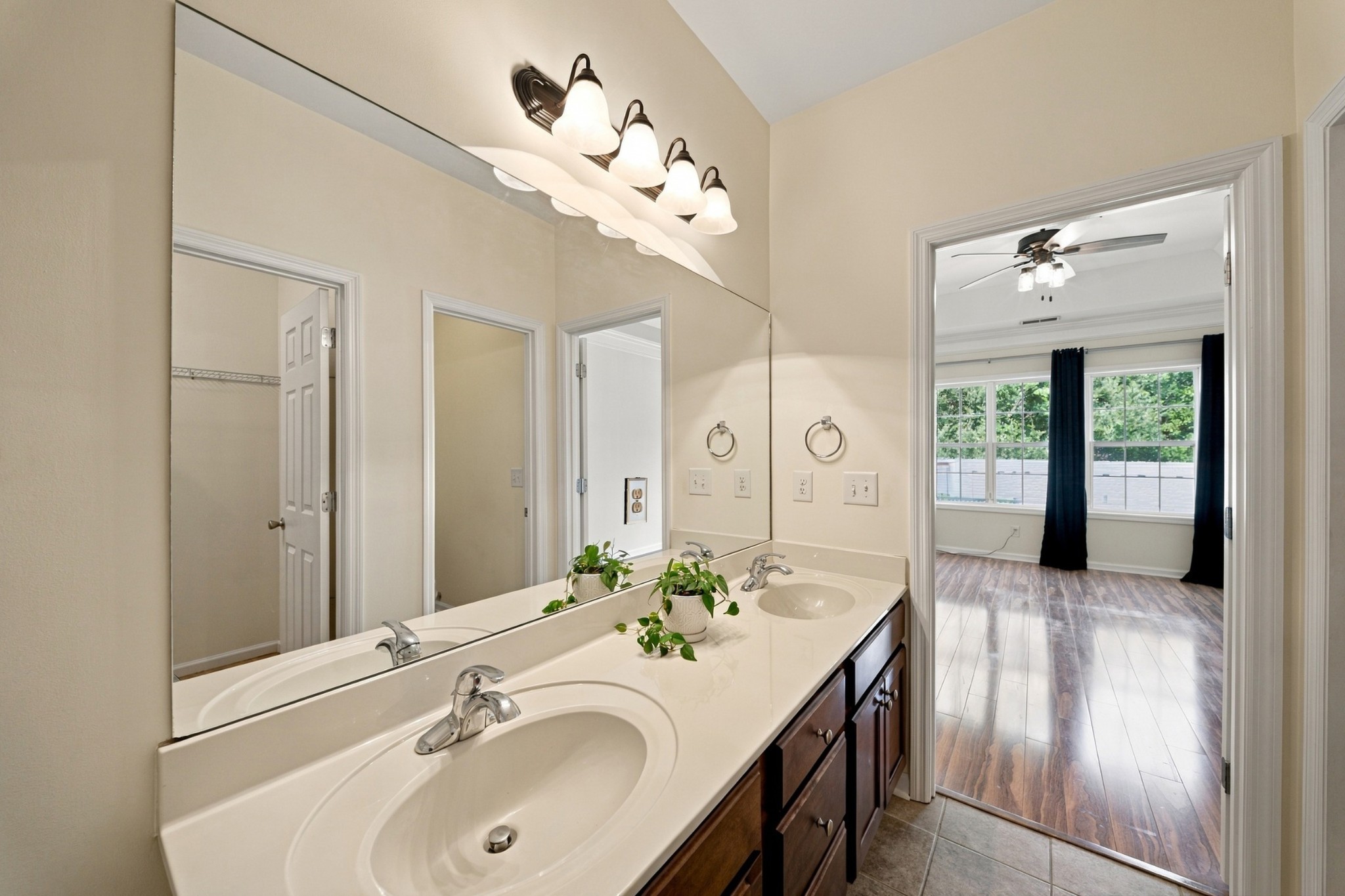 Bathroom with a double-sink vanity and large mirror, opening to a bright bedroom with wood-look flooring.