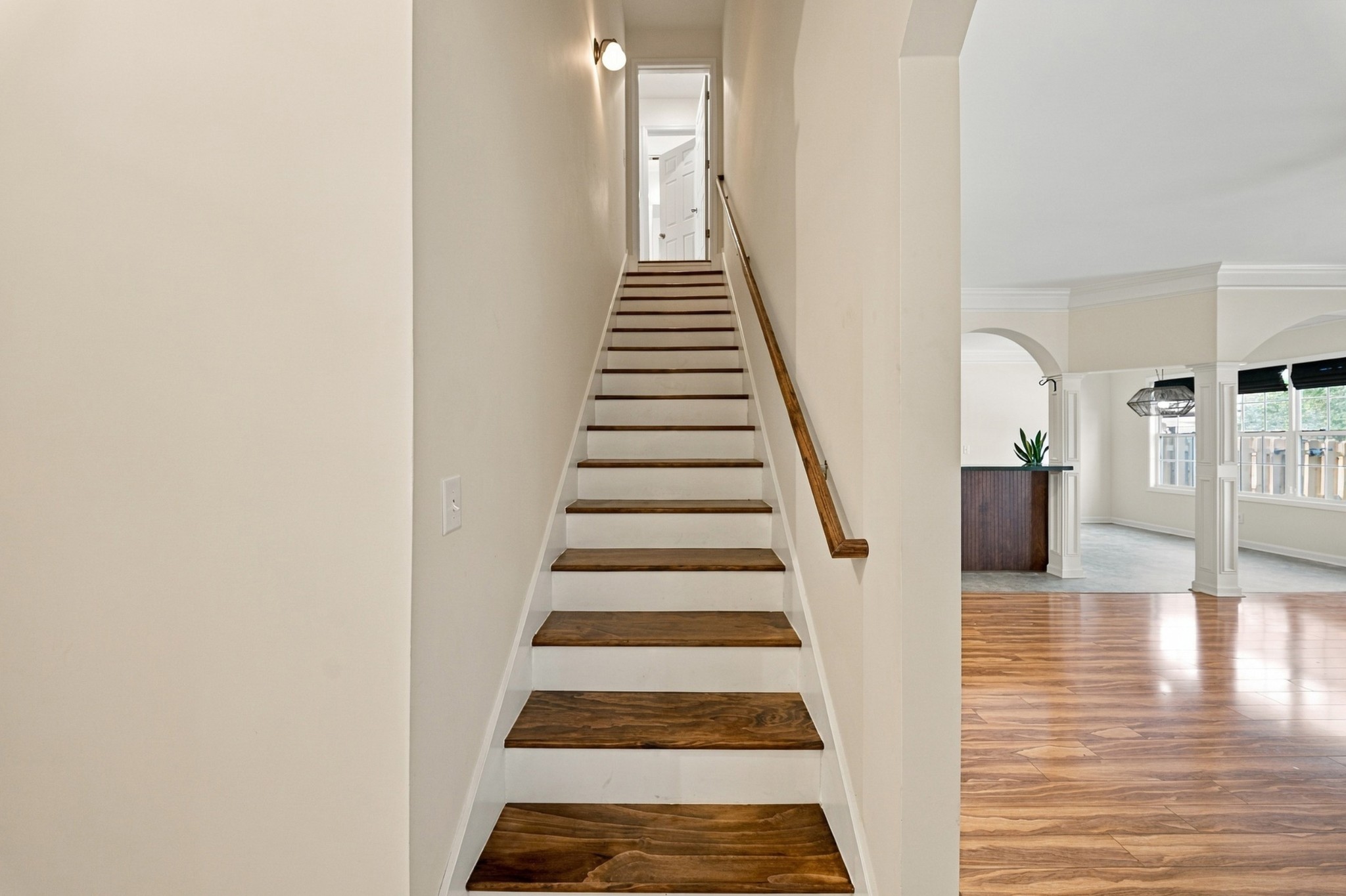 Staircase with dark wood treads and handrail beside a bright living area with hardwood floors.