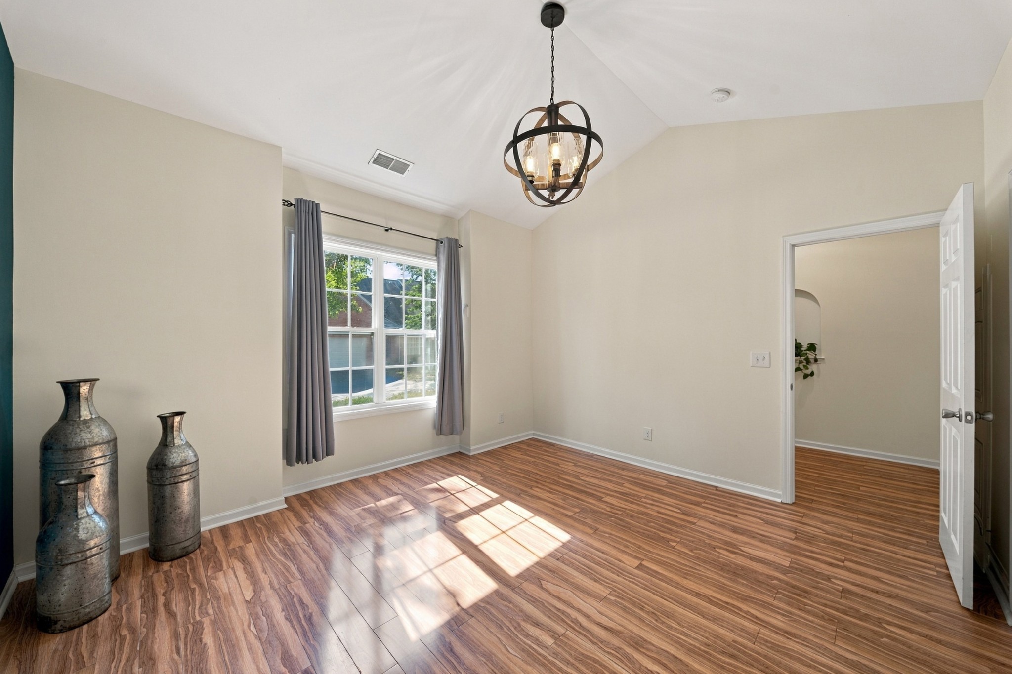 Empty bedroom with wood-look flooring, a vaulted ceiling, and sunlight streaming through a curtained window.