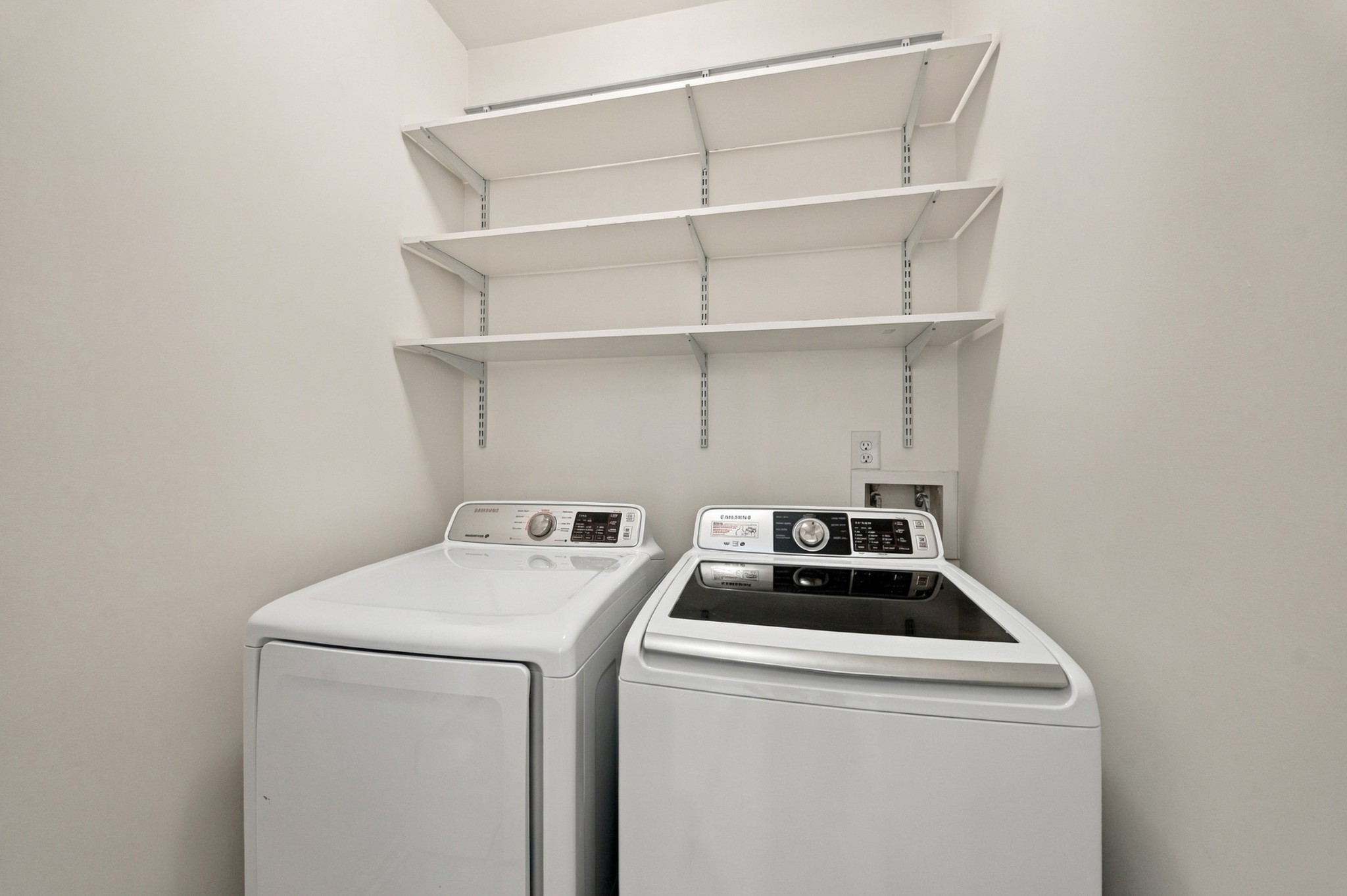 Laundry room with a white dryer and washer beneath three wall-mounted shelves.
