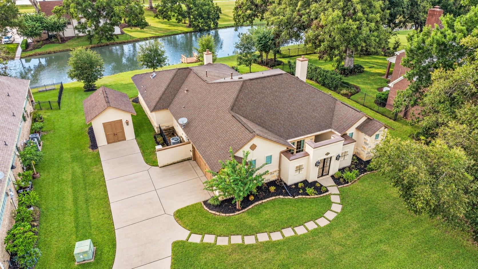 Aerial view of a cream home beside a tree-lined canal, with a broad driveway and detached outbuilding.