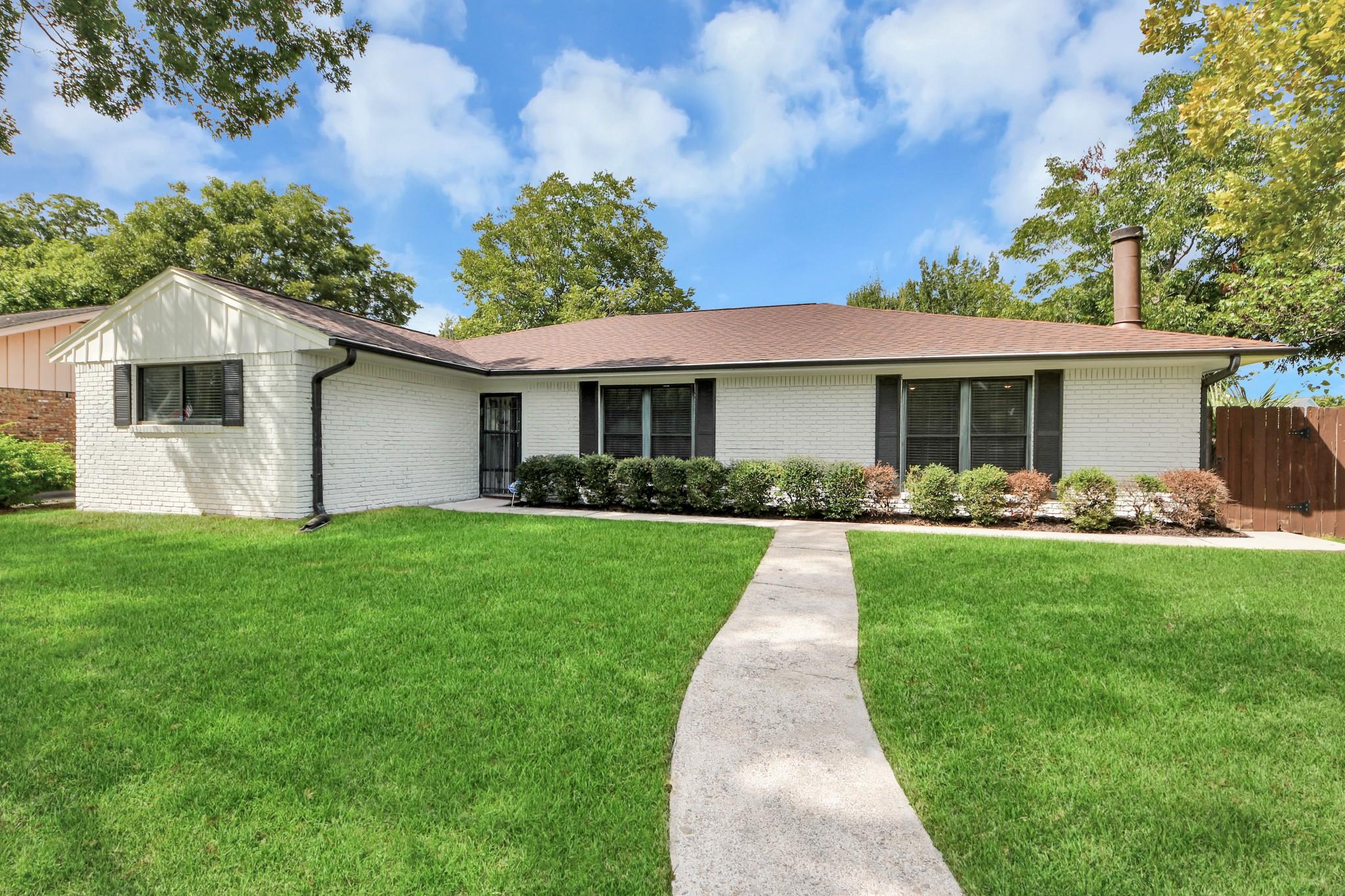 Single-story white brick house with dark shutters, a broad lawn, and a concrete walkway.