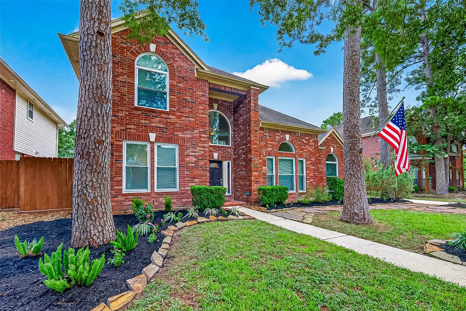 Two-story red-brick house with arched windows, landscaped beds, and an American flag.