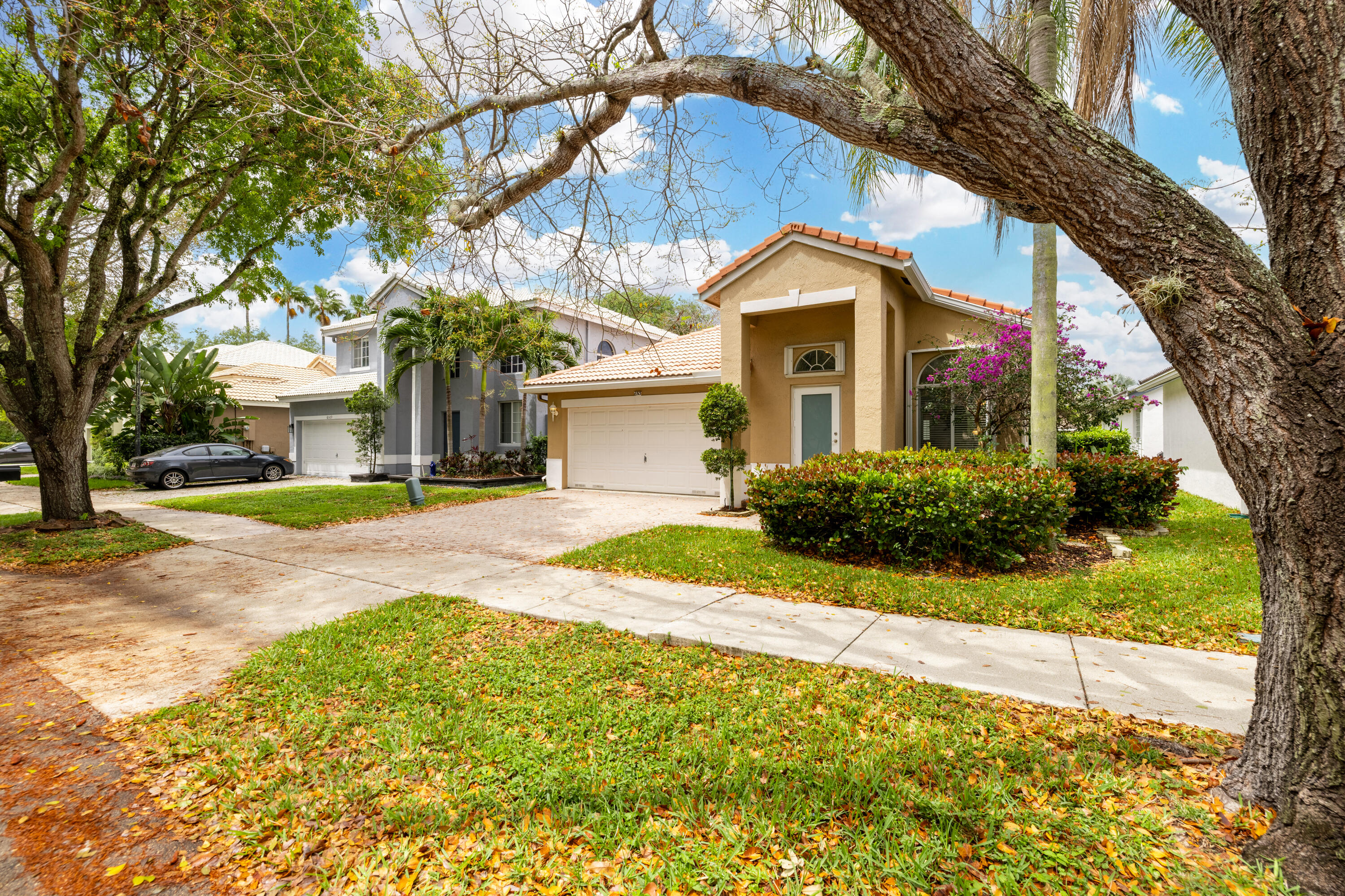 Tan stucco home with a tile roof and garage, framed by large trees and landscaped greenery.