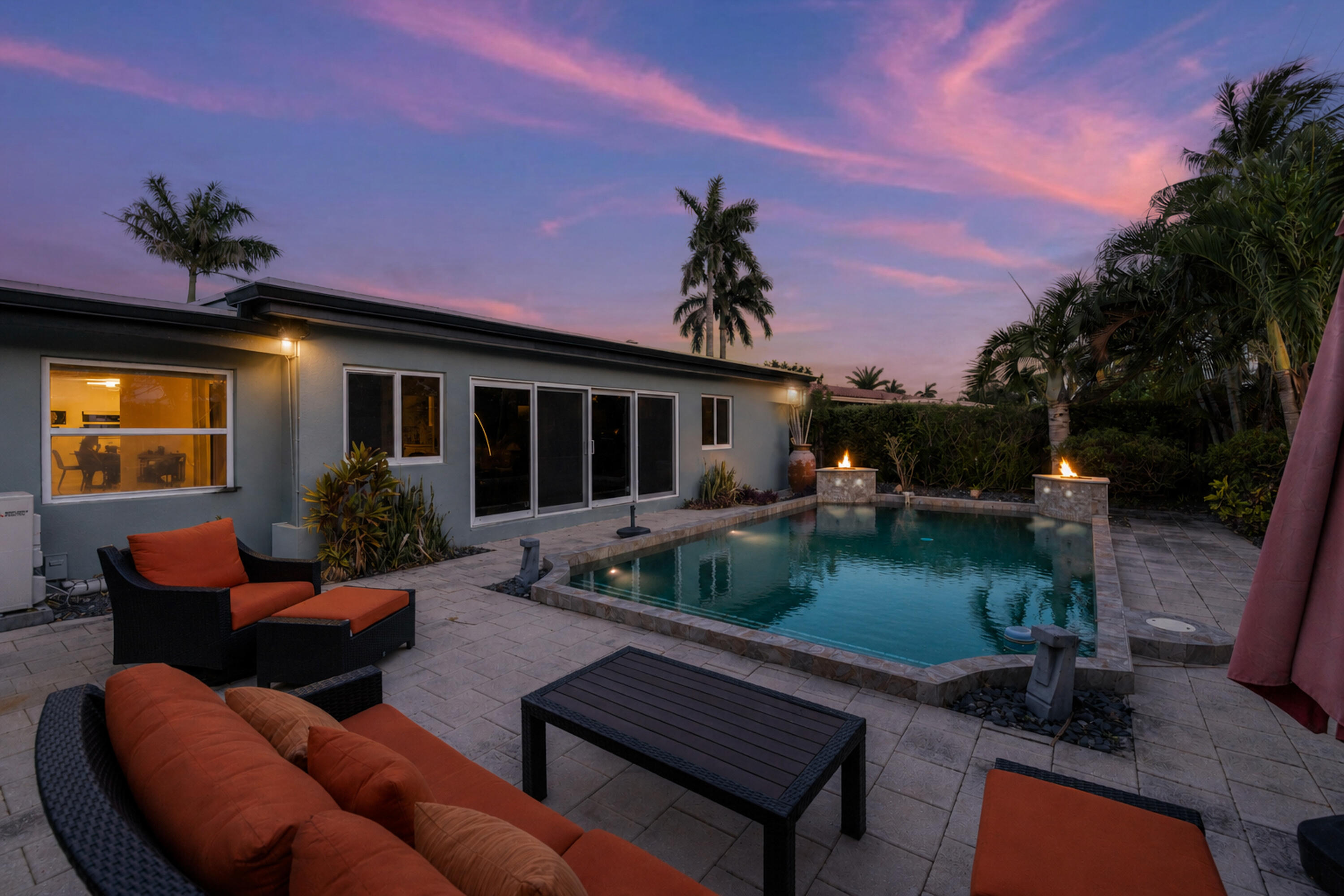 Backyard pool beside a single-story home, with patio seating and fire features beneath a pink dusk sky.