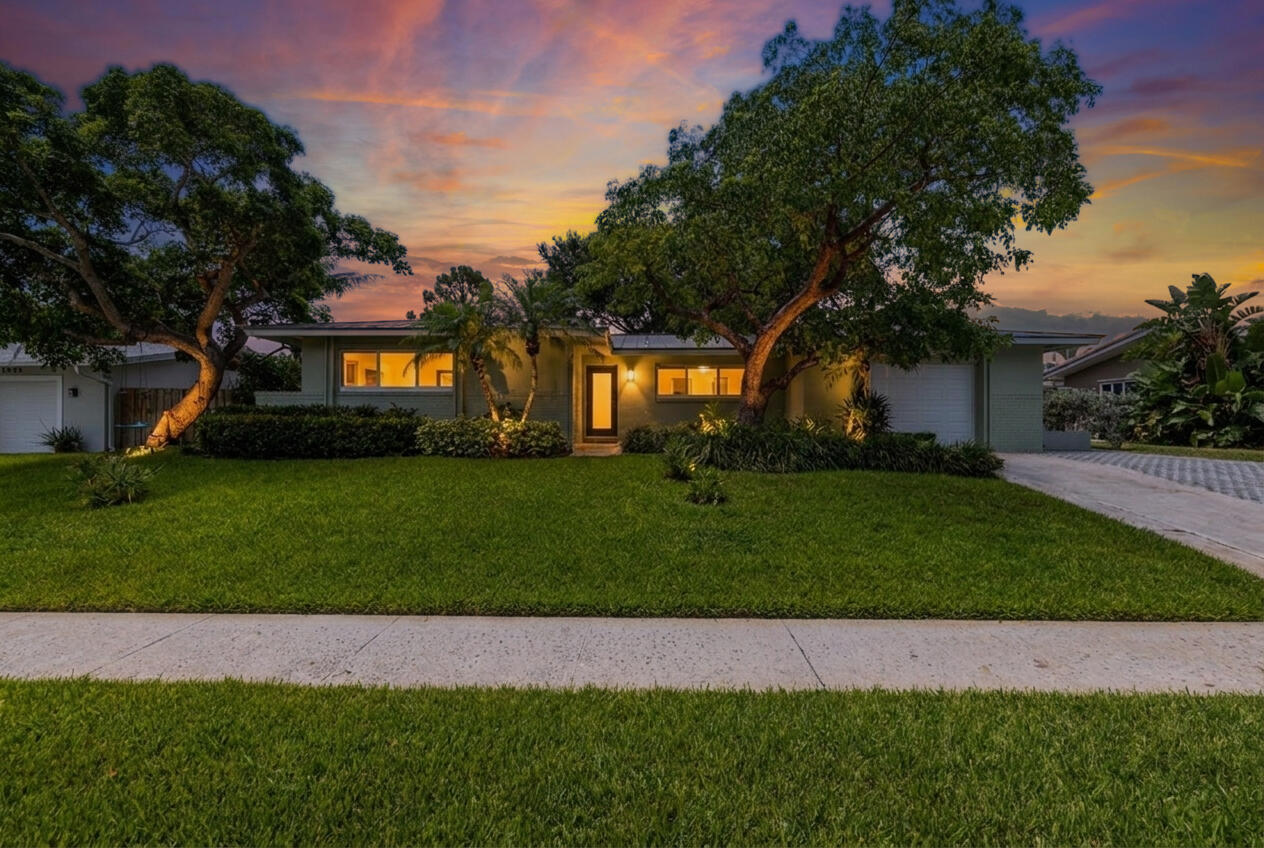 Single-story home at dusk, framed by mature trees, with lit windows and a broad front lawn.