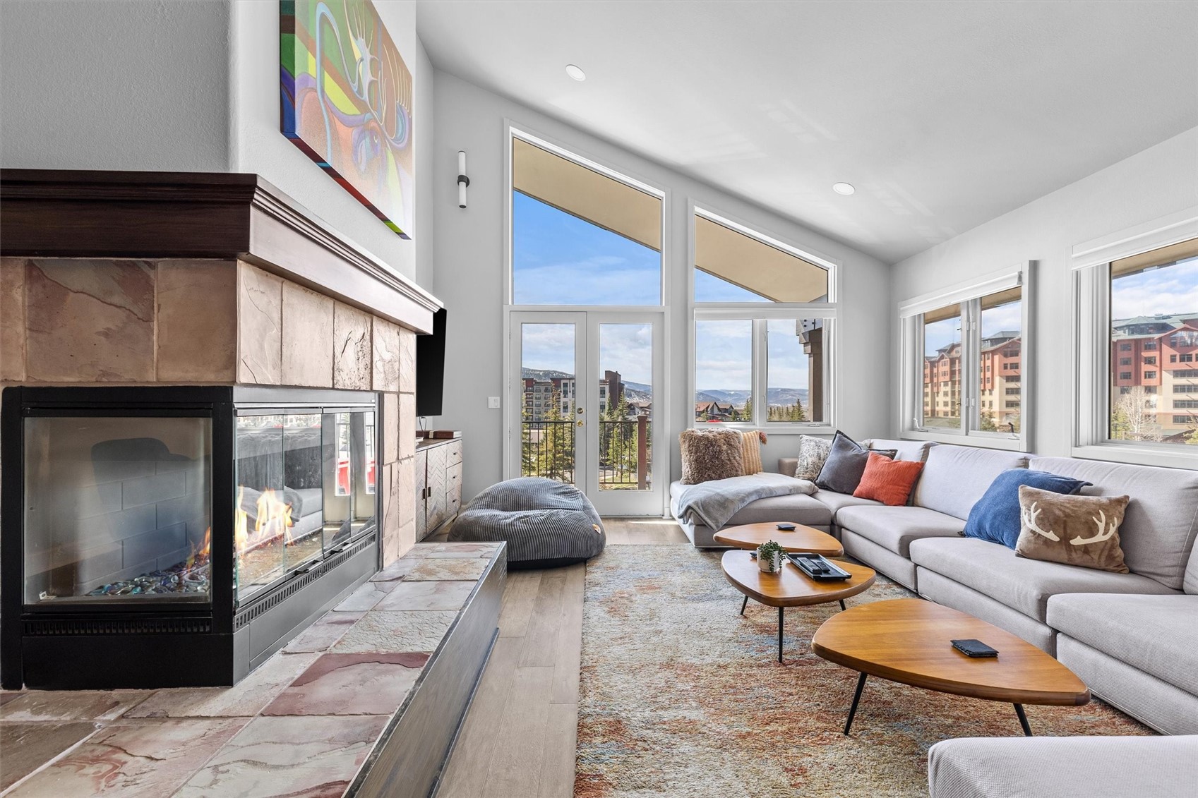 Living room with a stone fireplace, gray sectional, and tall windows overlooking mountains.