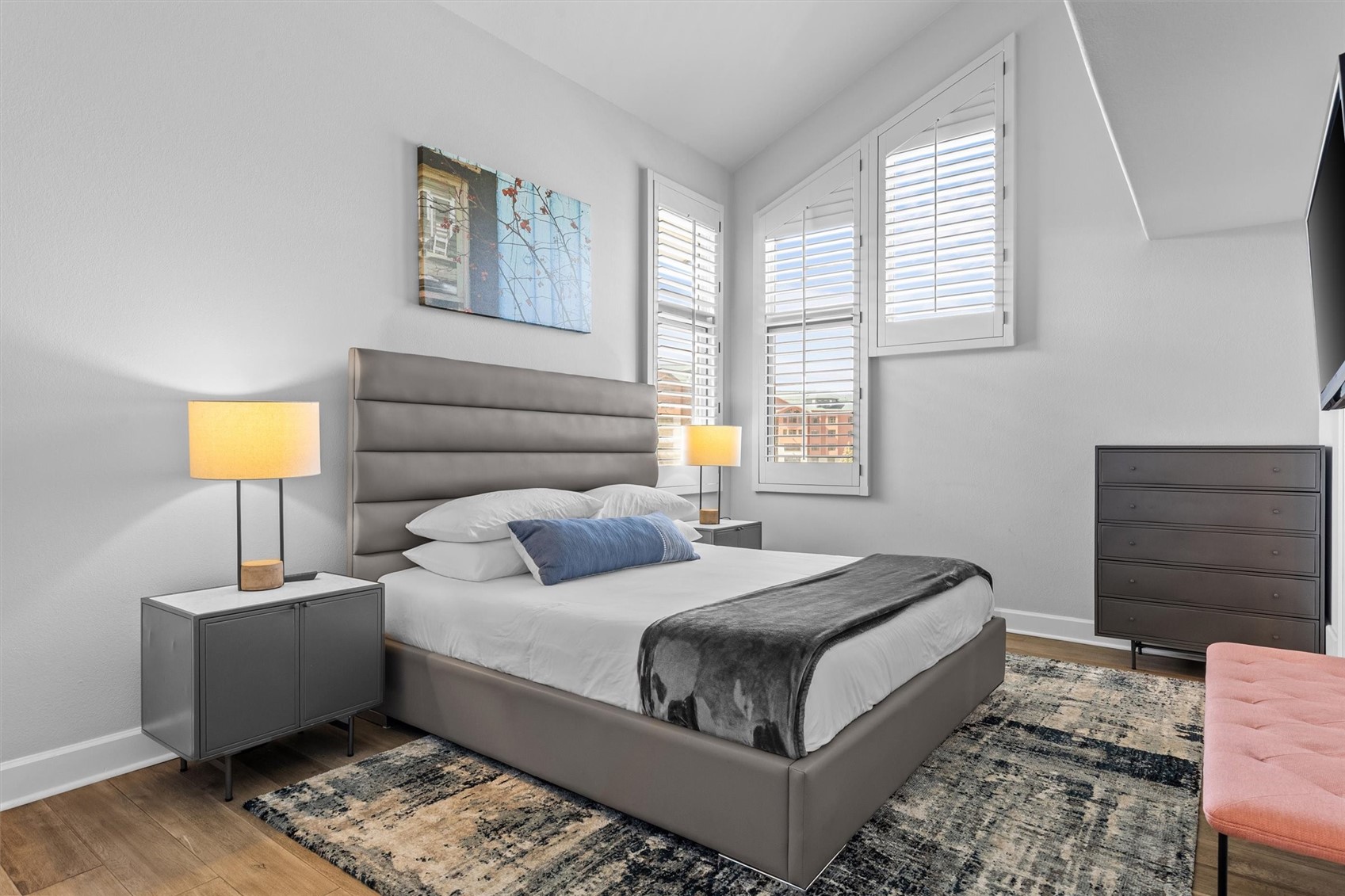 Furnished bedroom with a gray upholstered bed, shuttered windows, and a colorful area rug.