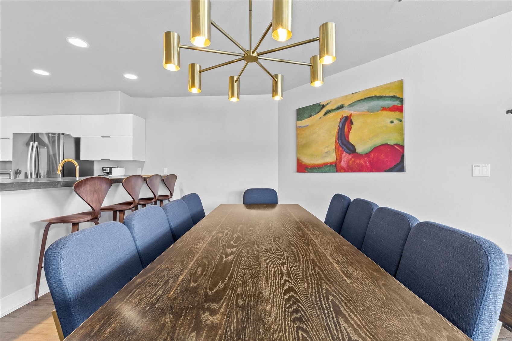 Dining room with a long wood table, blue chairs, and a brass chandelier beside an open kitchen.