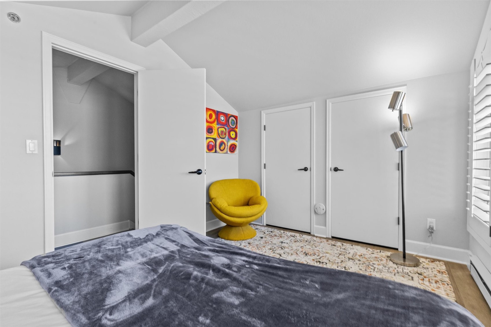 Bedroom with a gray-covered bed, yellow accent chair, colorful art, and white shutters.