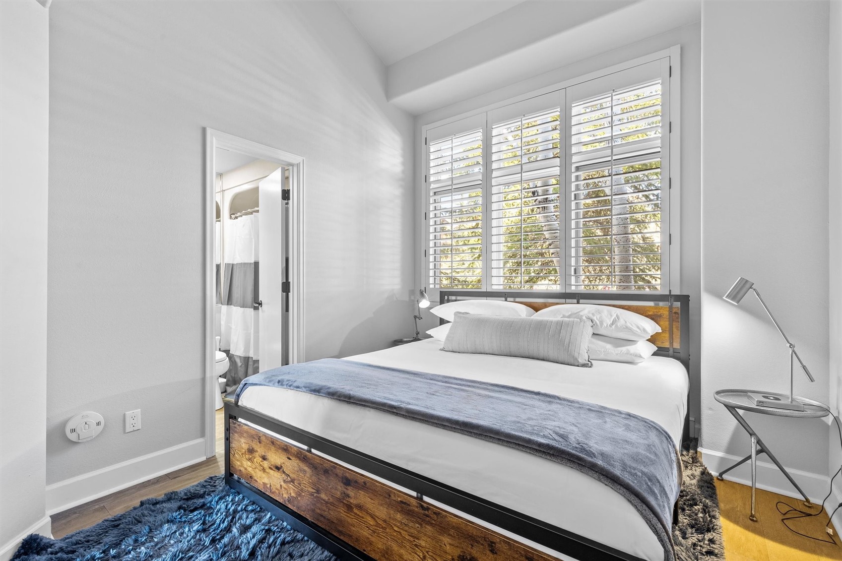 Bedroom with a wood-and-metal bed, blue-gray throw, plantation shutters, and adjoining bathroom doorway.