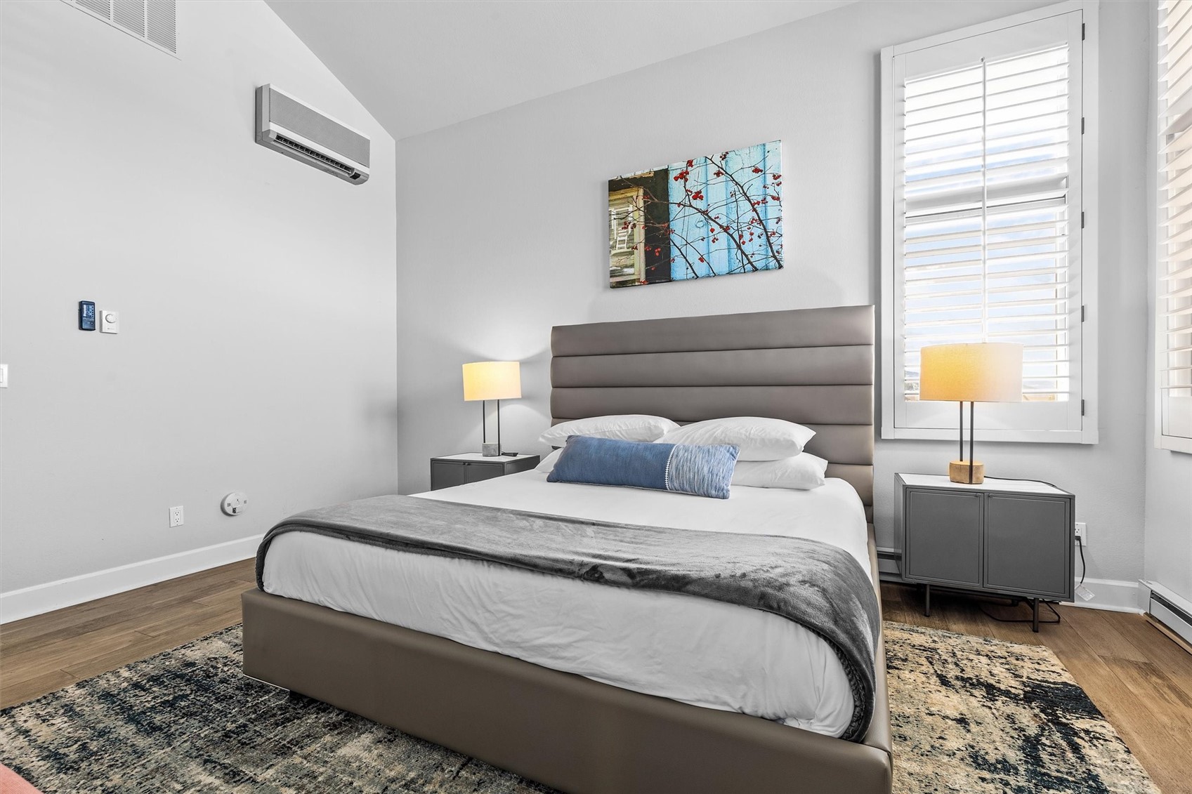 Bedroom with a large gray upholstered bed, matching nightstands, and white plantation shutters.