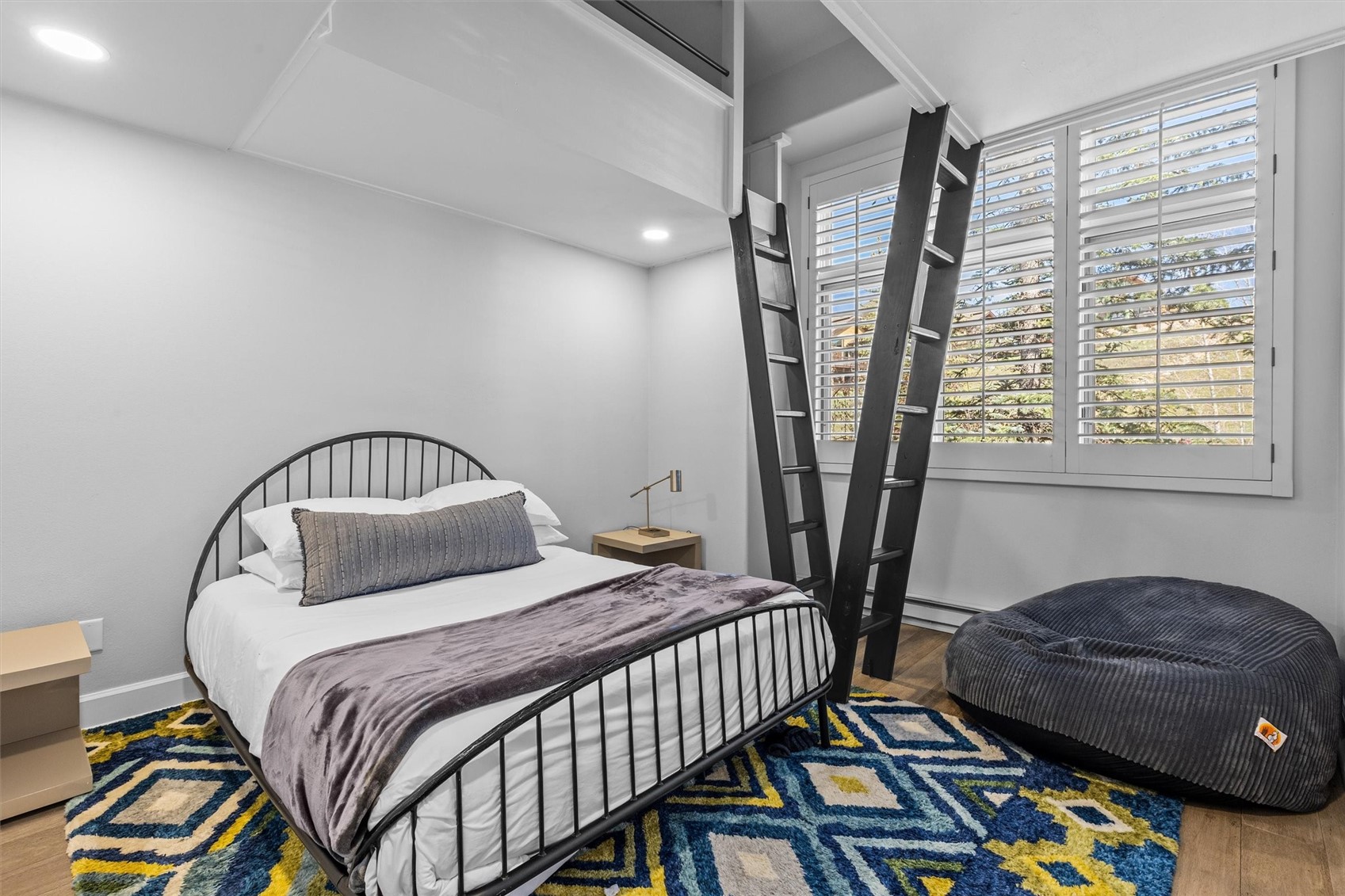 Bedroom with a metal bed, overhead loft ladder, shuttered windows, and a large round floor cushion.