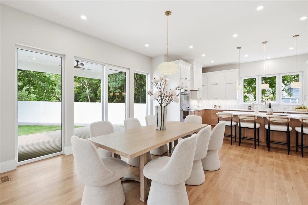 Open dining room and kitchen with a light-wood table, white cabinetry, and large garden-facing windows.