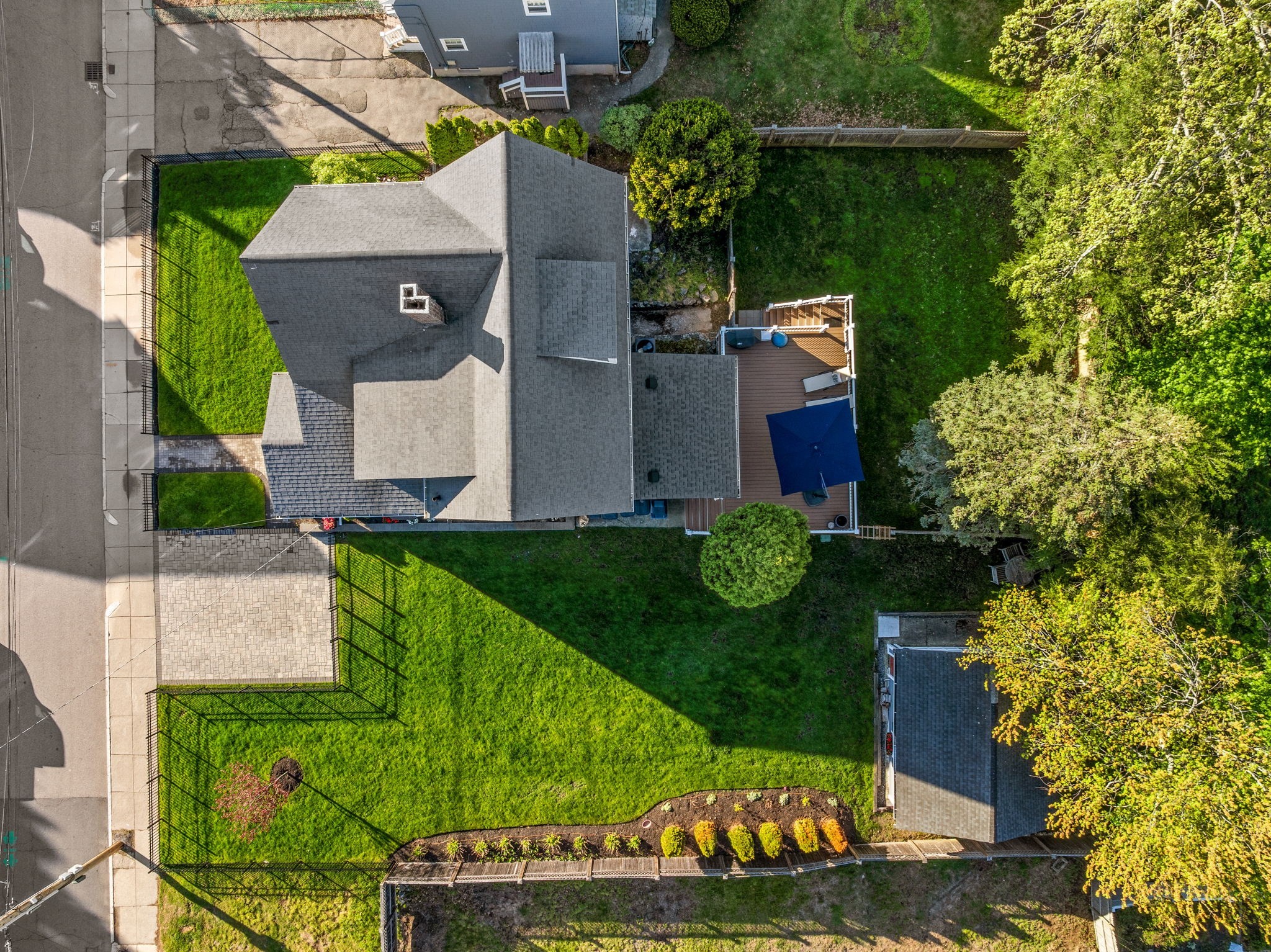Aerial view of a house with a fenced green lawn, paved driveway, and rear deck with a blue canopy.