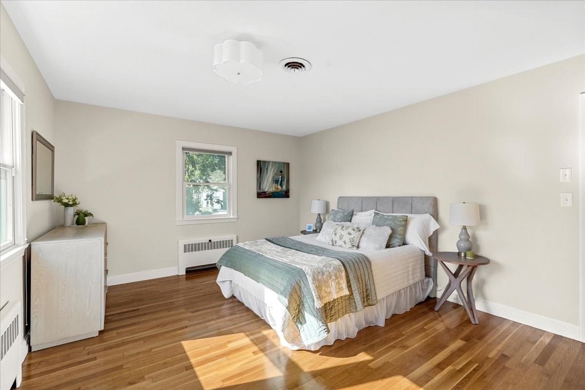 Furnished bedroom with a gray upholstered bed, layered bedding, and sunlit hardwood floors.