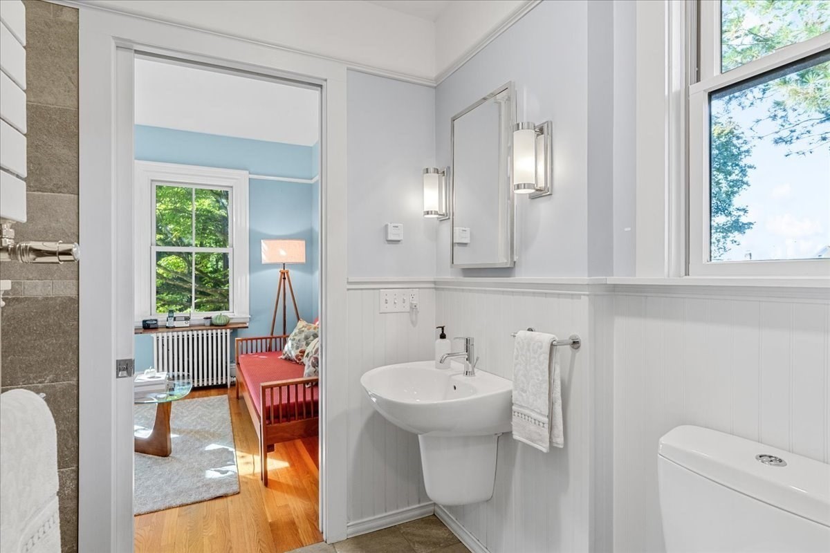 Bright bathroom with a pedestal sink and large window, looking through to a blue bedroom with a red bed.