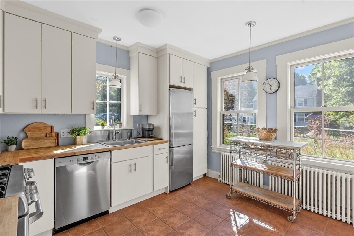 Kitchen with white cabinets, butcher-block counters, stainless appliances, and a rolling cart by large windows.