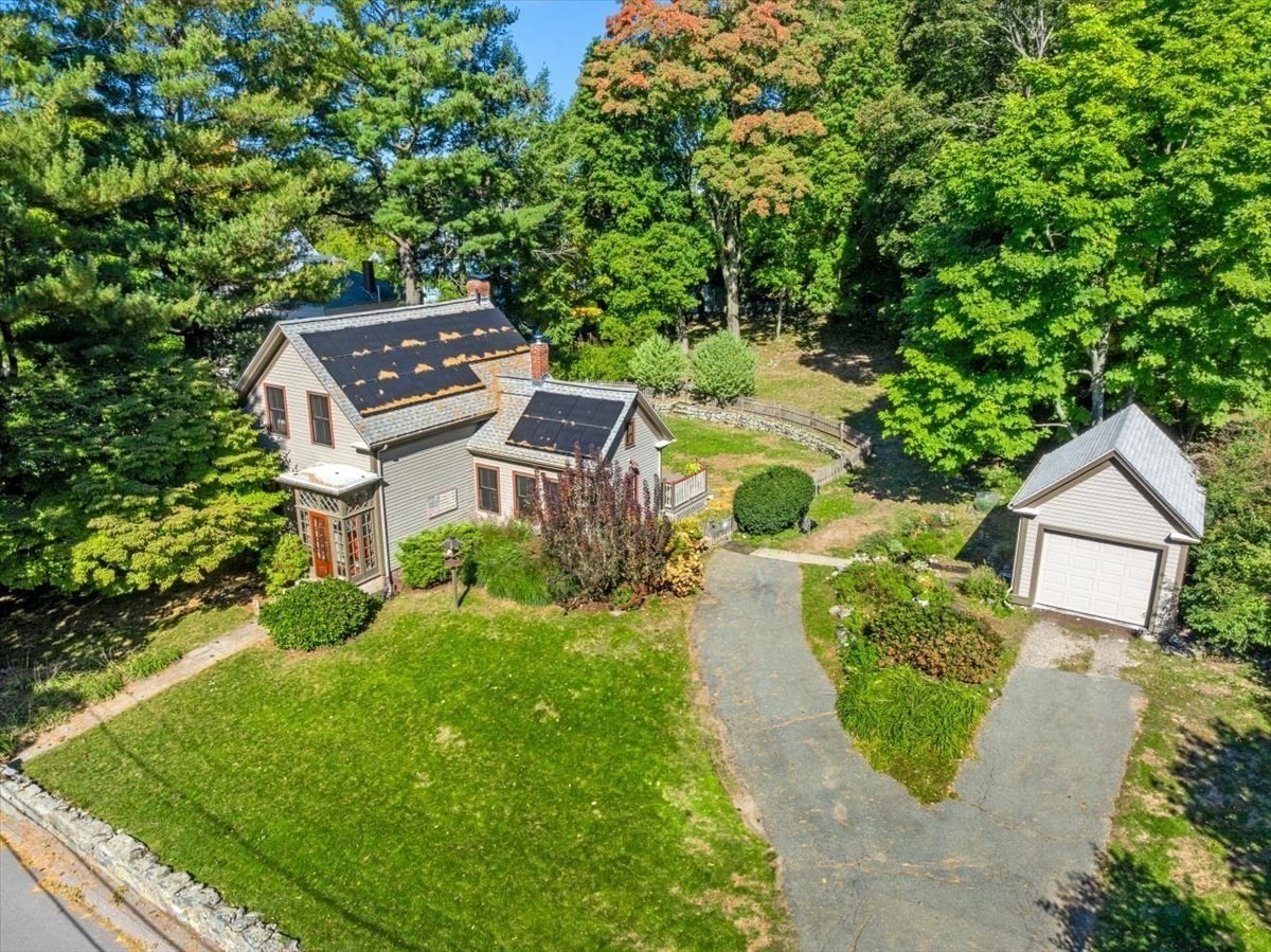 Aerial view of a gray house with rooftop solar panels, detached garage, and tree-lined lawn.