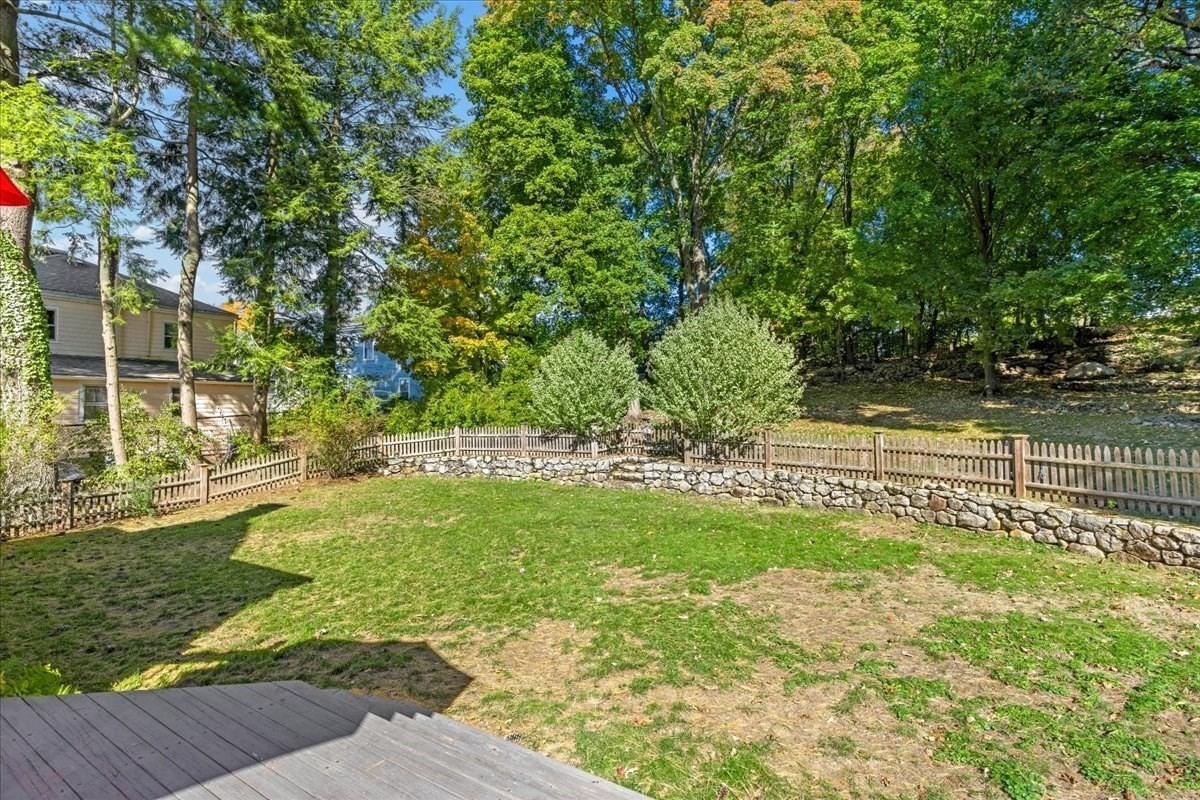 Backyard lawn with a wooden deck, curved stone wall, picket fence, and mature trees.