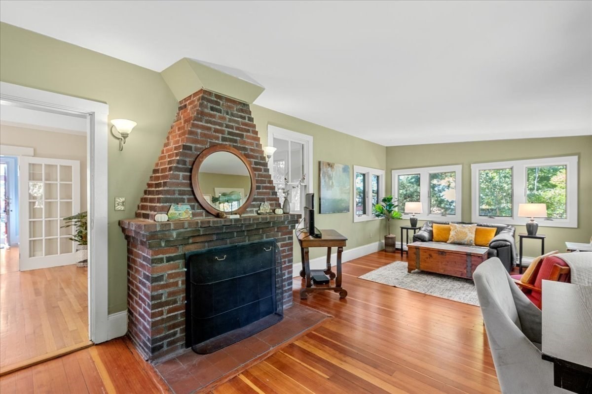 Living room with a brick fireplace, round mirror, hardwood floors, and a gray sofa by bright windows.