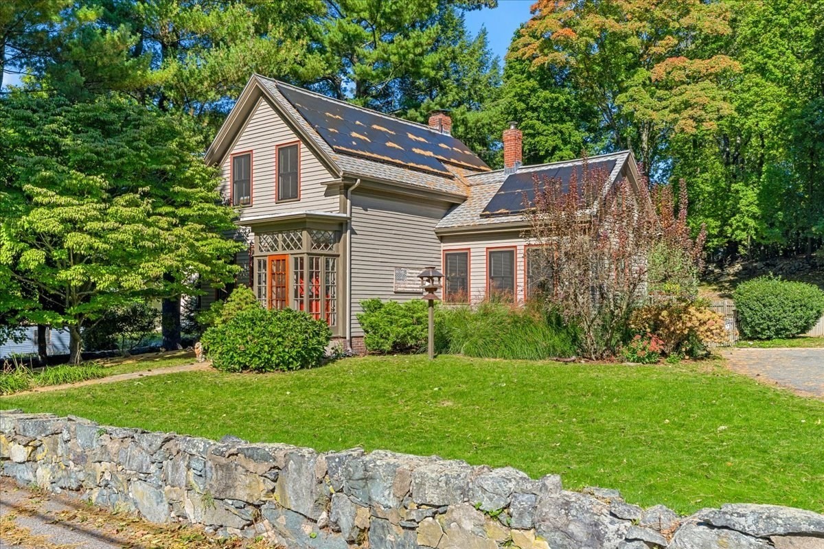 Gray two-story house with roof solar panels, surrounded by mature trees behind a stone wall.