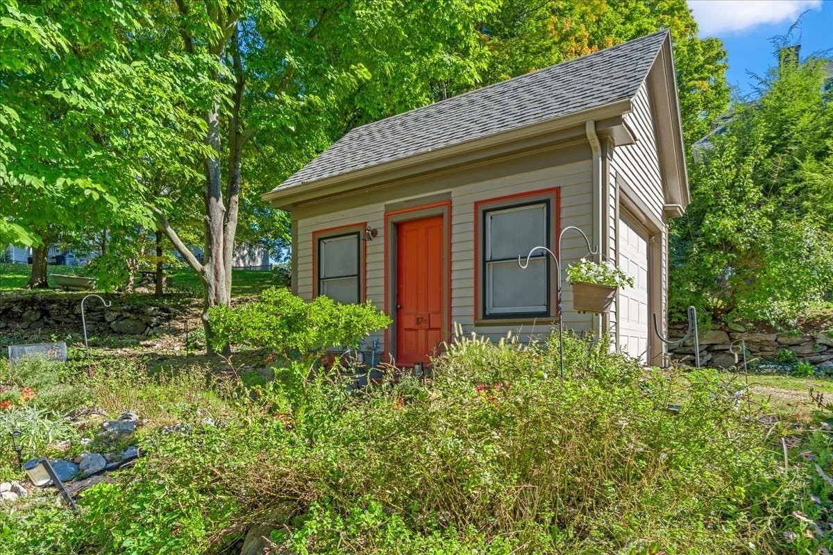 Small gray-sided structure with a red-orange door, nestled among leafy trees and garden shrubs.