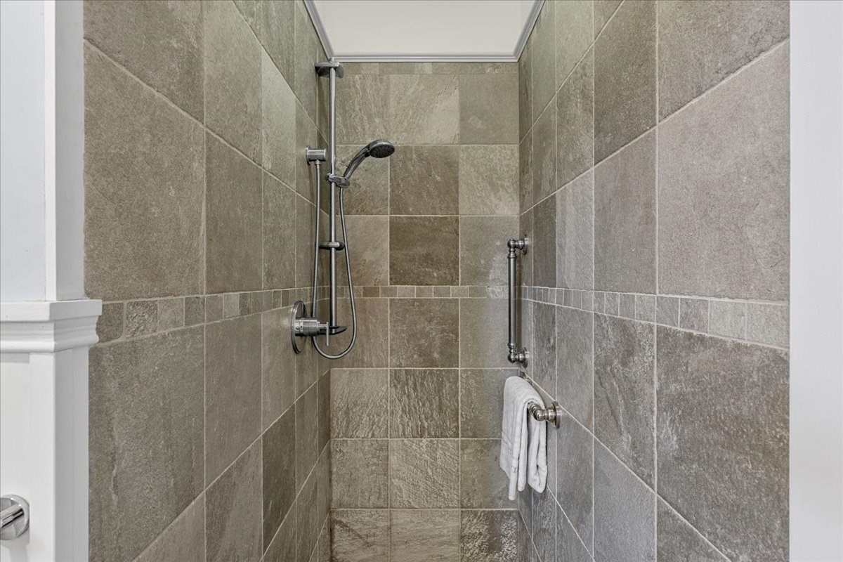 Walk-in shower with gray tile, a handheld showerhead, chrome grab bar, and a white towel.
