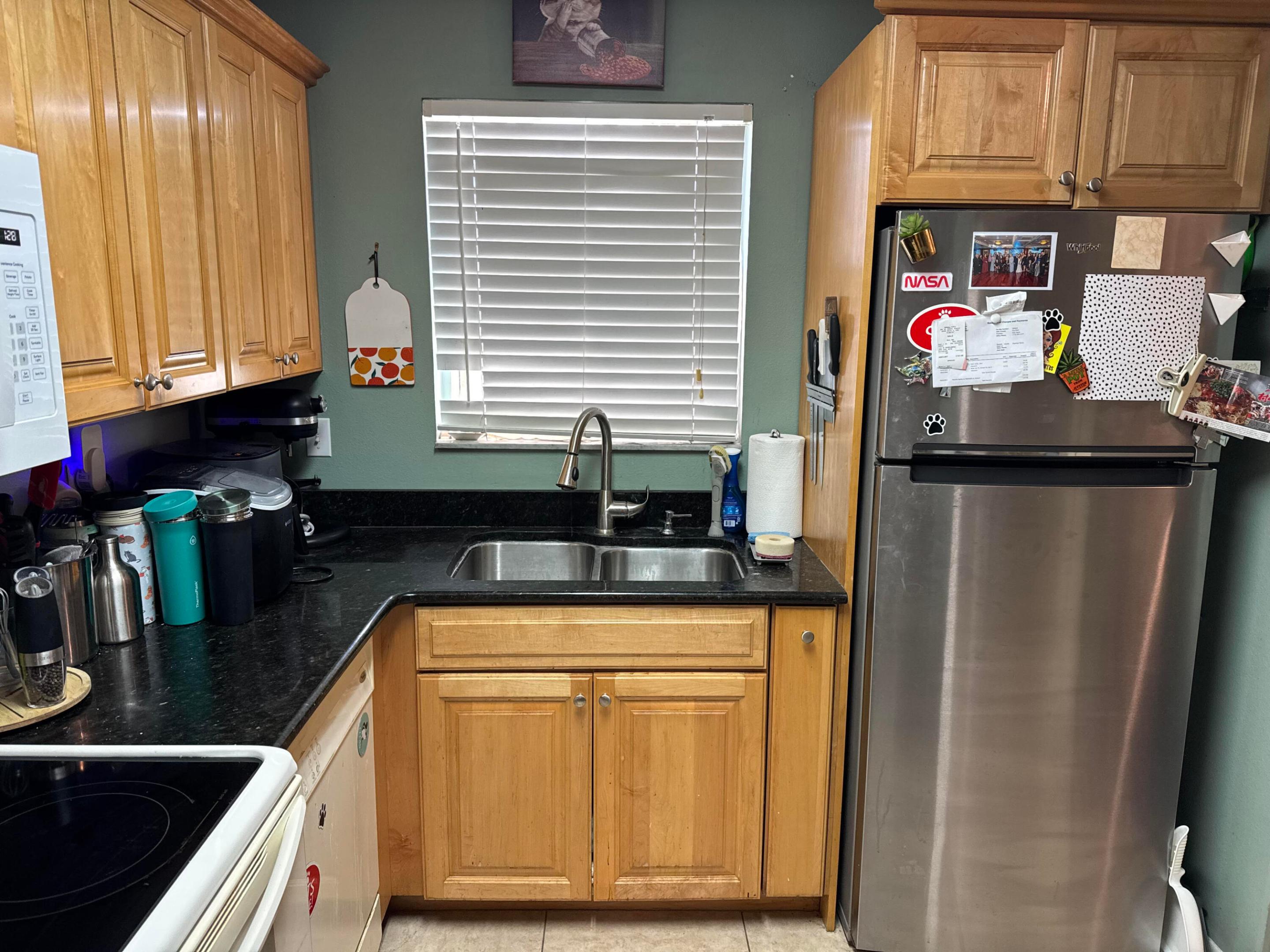Kitchen with honey-colored wood cabinets, black countertops, and a double sink beside a magnet-covered stainless-steel refrigerator.