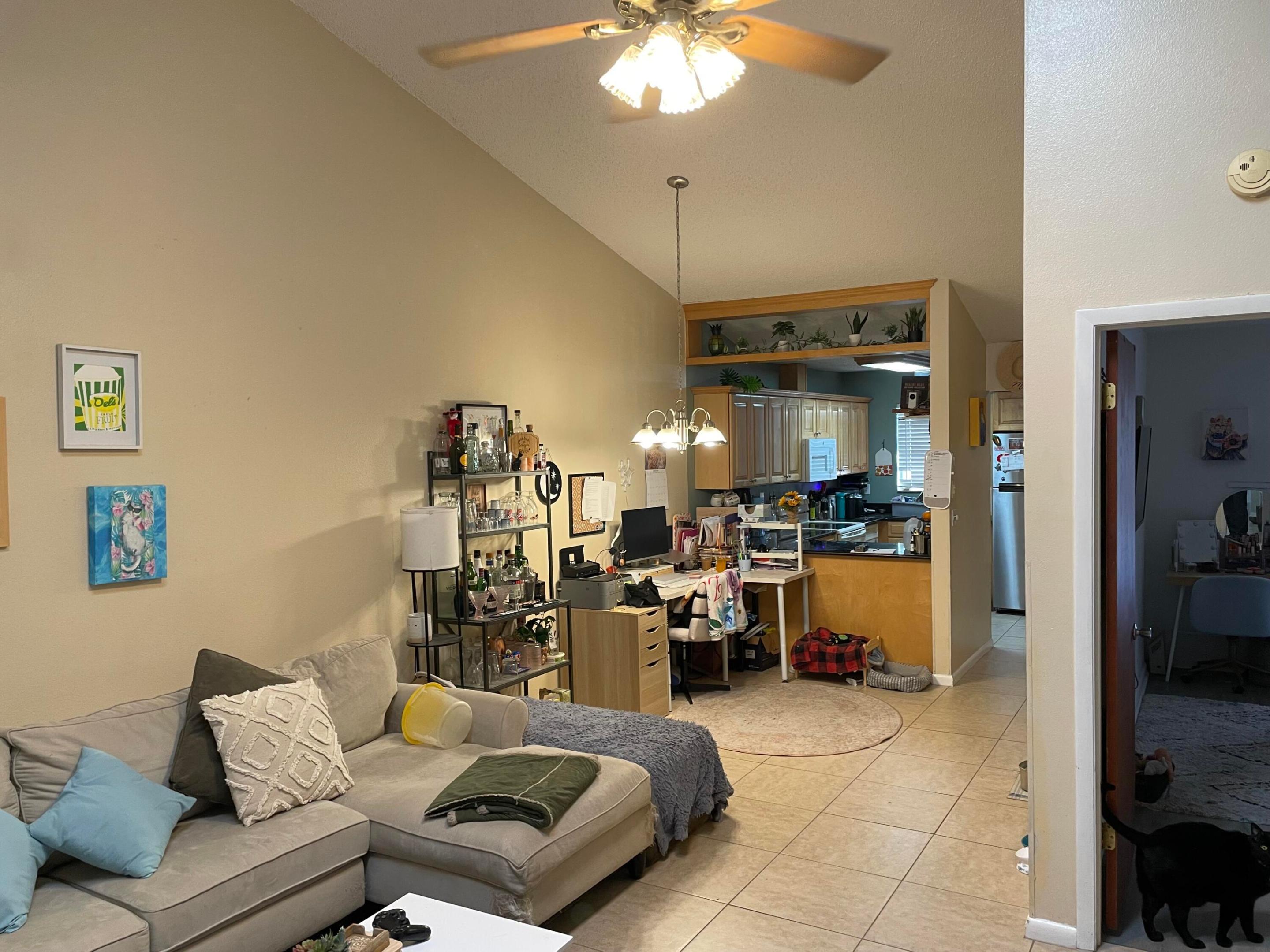 Living room with a beige sectional opening to a kitchen, with tiled floors and a ceiling fan.