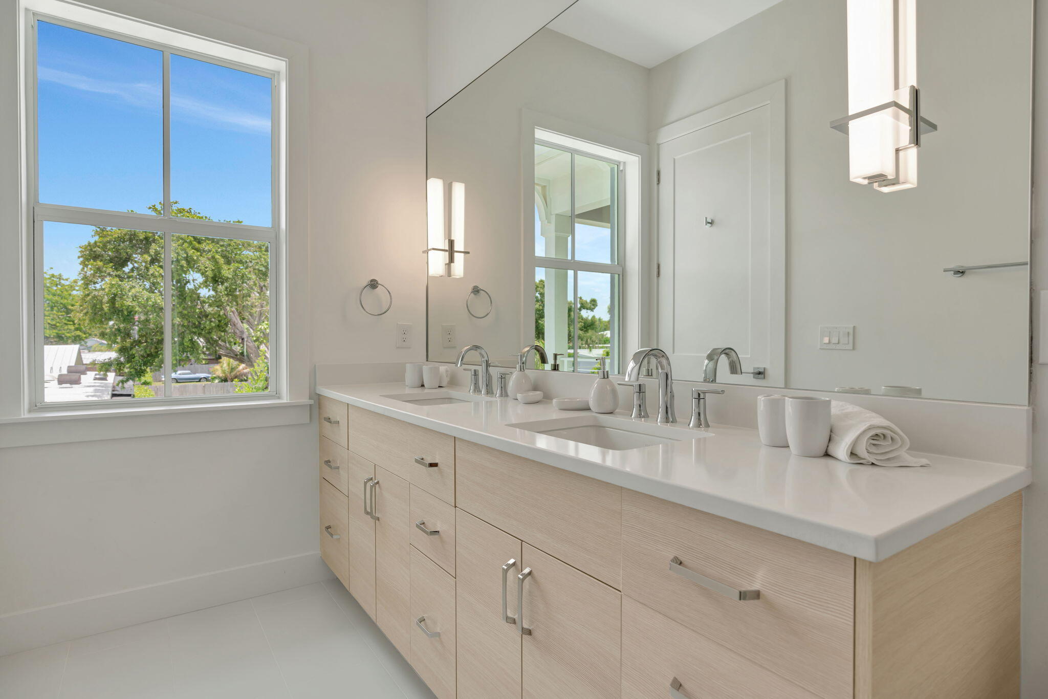 Bright bathroom with a double-sink vanity, pale wood cabinets, and a large window overlooking trees.