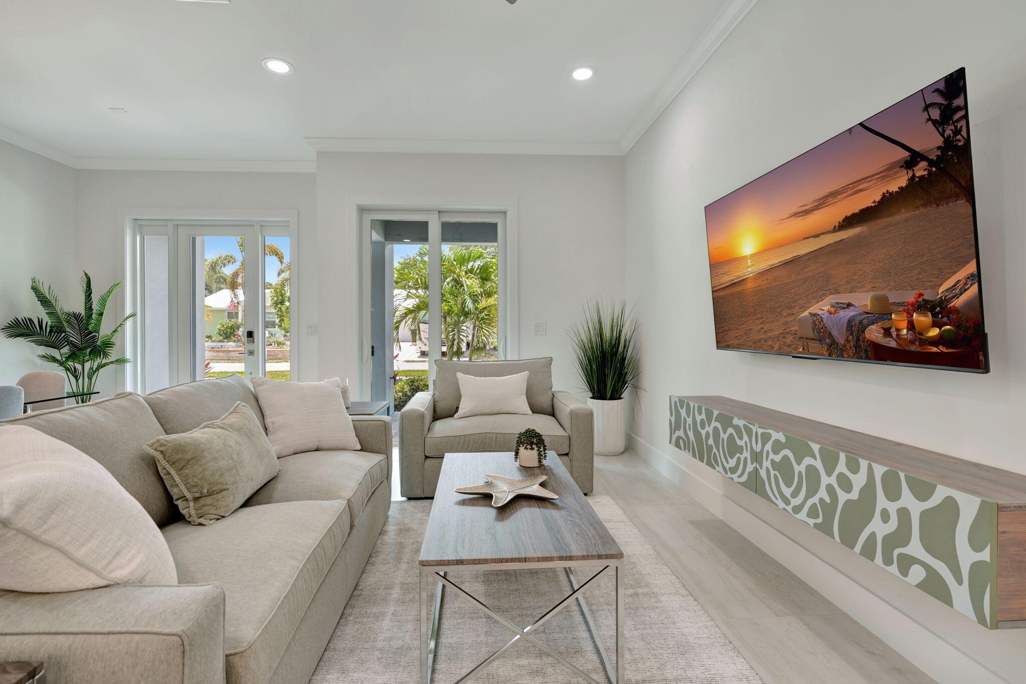 Furnished living room with a neutral sectional, glass doors, and a wall-mounted screen showing a beach sunset.