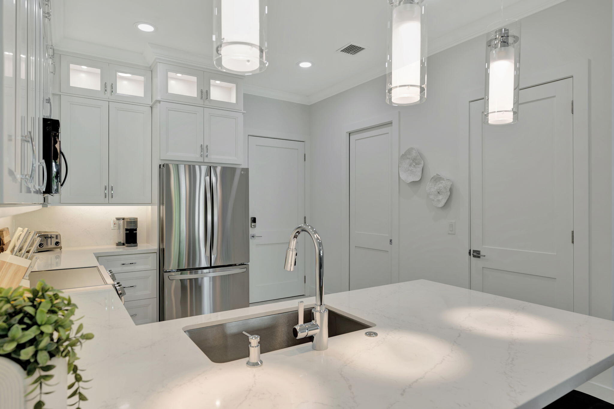 Modern white kitchen with a quartz island, undermount sink, stainless steel refrigerator, and glass pendant lights.