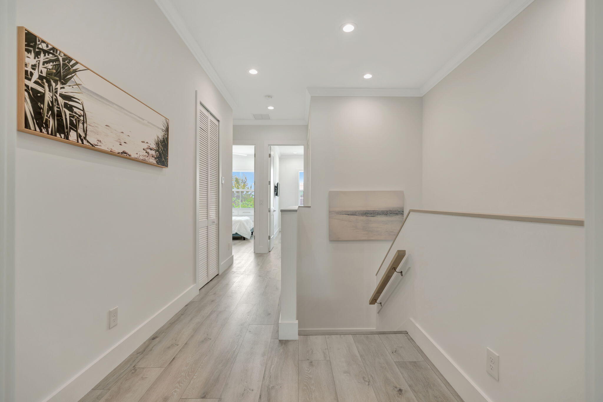 Bright upstairs hallway with light flooring, coastal artwork, and a stair landing beside an open bedroom doorway.